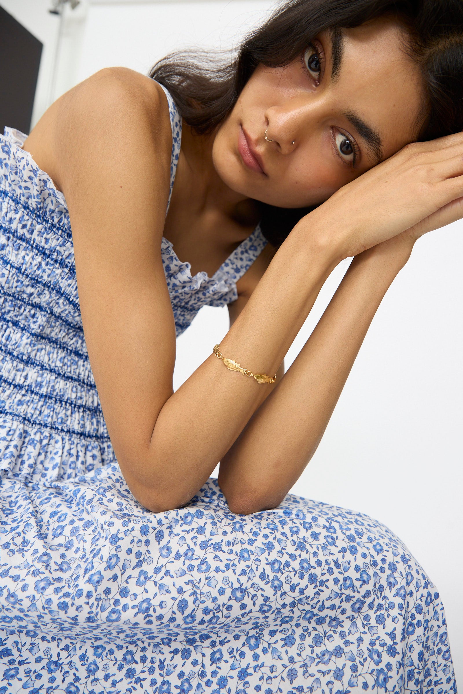 A woman in a blue and white floral dress sits with her arms on her knees, looking at the camera. She wears the Grainne Morton Fish Bracelet and has long dark hair.