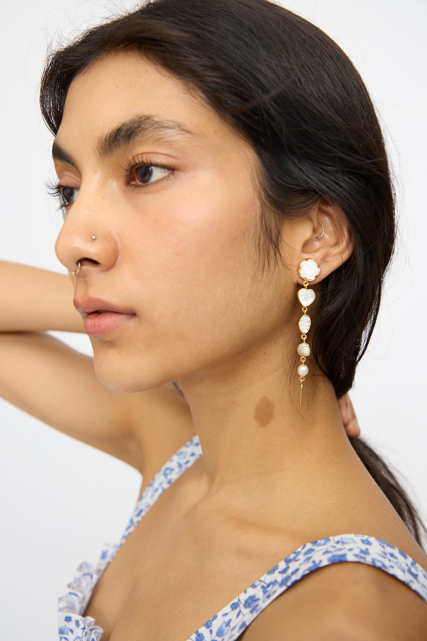 A woman with long dark hair, a floral dress, nose stud, and a visible neck birthmark wears Grainne Morton Five Charm with Victorian Drop Earrings, featuring 18K gold plating, pearls, and heart details.