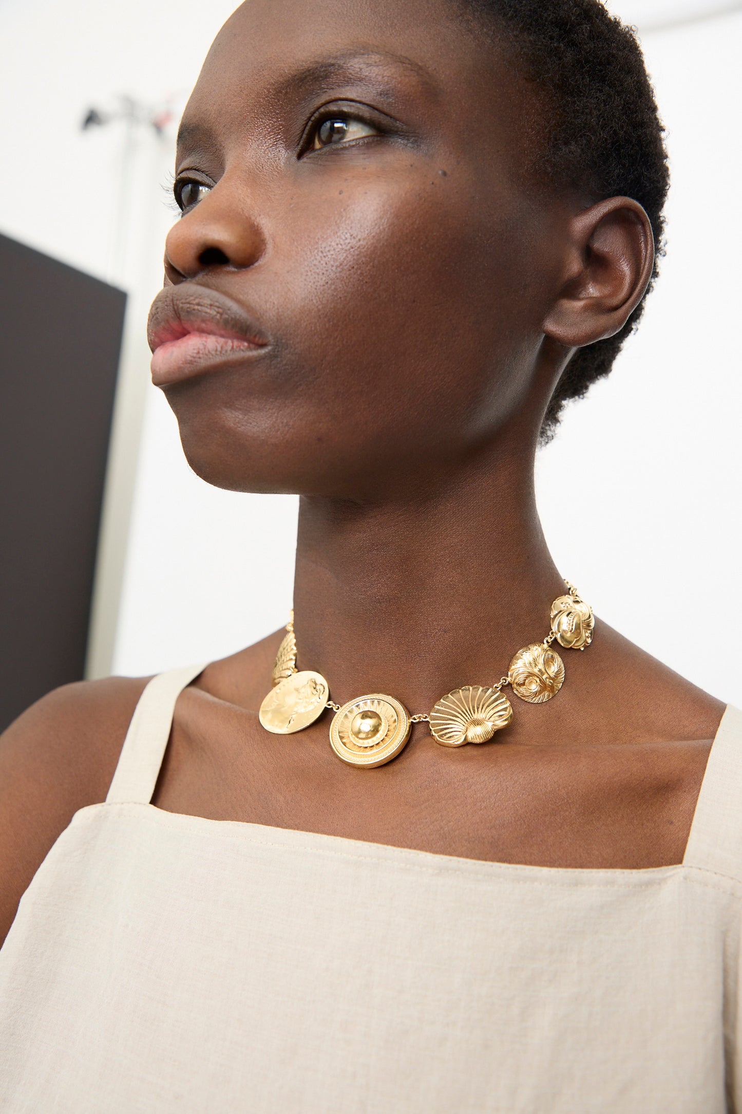 A person wearing a beige sleeveless top and the Grainne Morton Gold Button Necklace, a chunky gold plated silver belcher chain with ornate charms, looking slightly to the side.