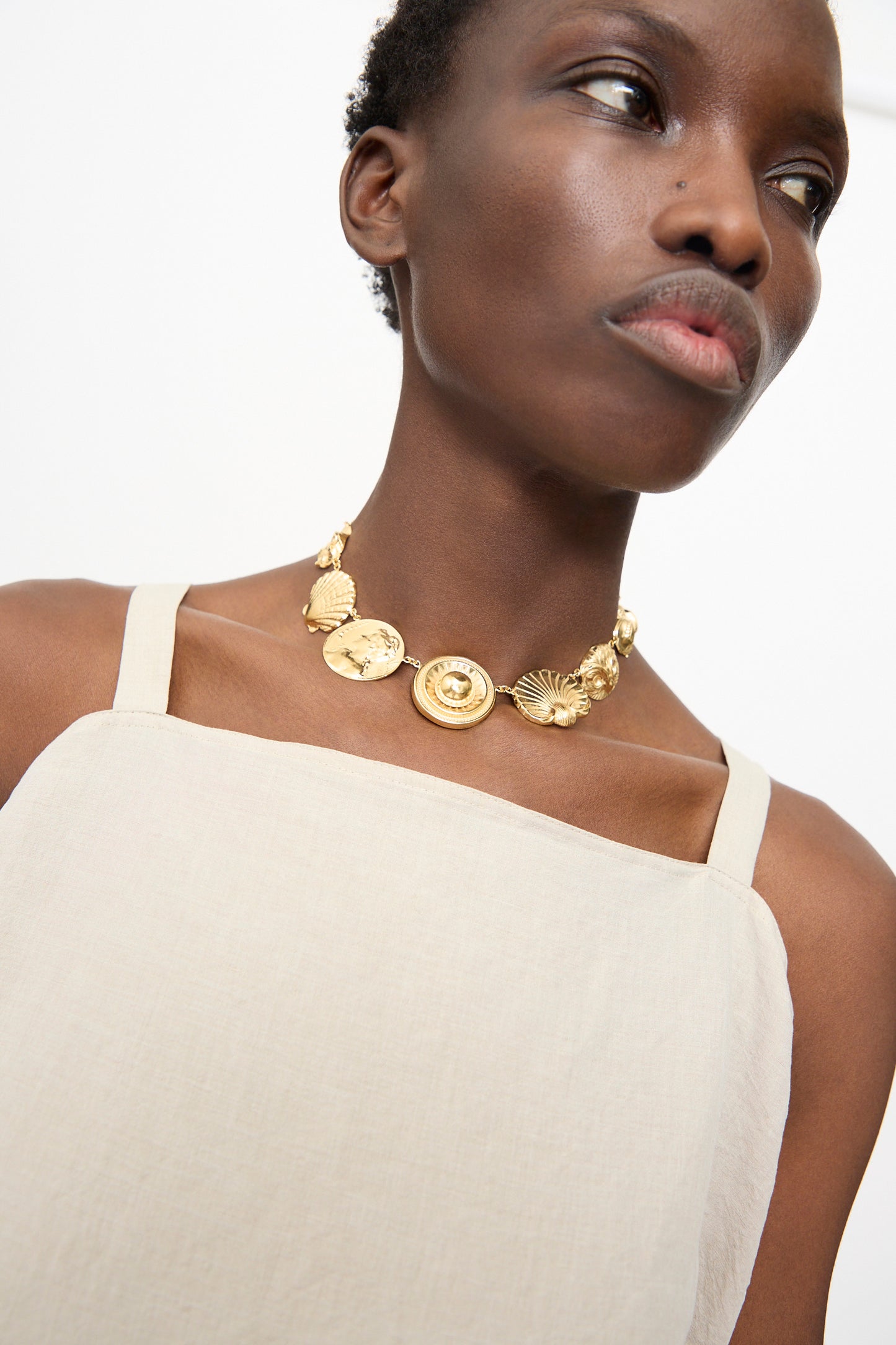 A person wearing a beige square-neck top and the Grainne Morton Gold Button Necklace, looking slightly away from the camera.