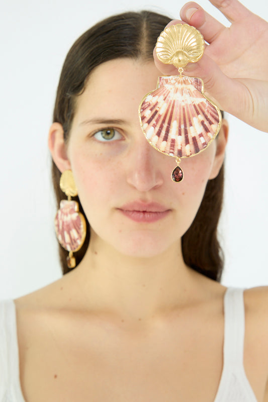 A woman with long brown hair wears a white tank top and holds a Grainne Morton Scallop Earring against her face, showcasing the matching earring against a plain background.