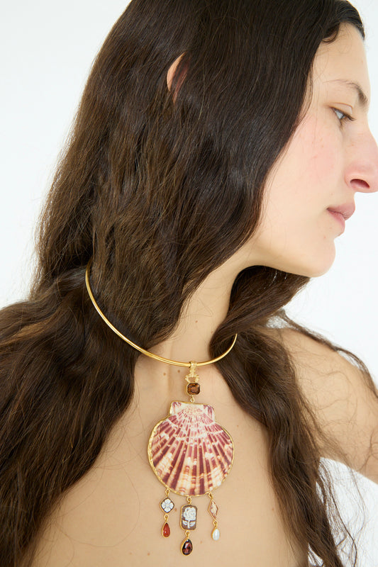 A person with long dark hair wears the stunning Grainne Morton Scallop Necklace, handcrafted in Edinburgh. The 18k gold-plated silver piece showcases a large seashell and dangling gemstone ornaments, adding an air of elegance.