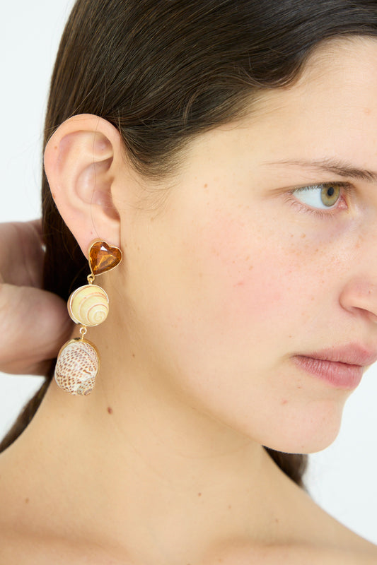 A woman wearing Grainne Morton's Shell Charm Drop Earrings, featuring a heart-shaped stone and two shell pieces, crafted from 18k gold-plated silver.