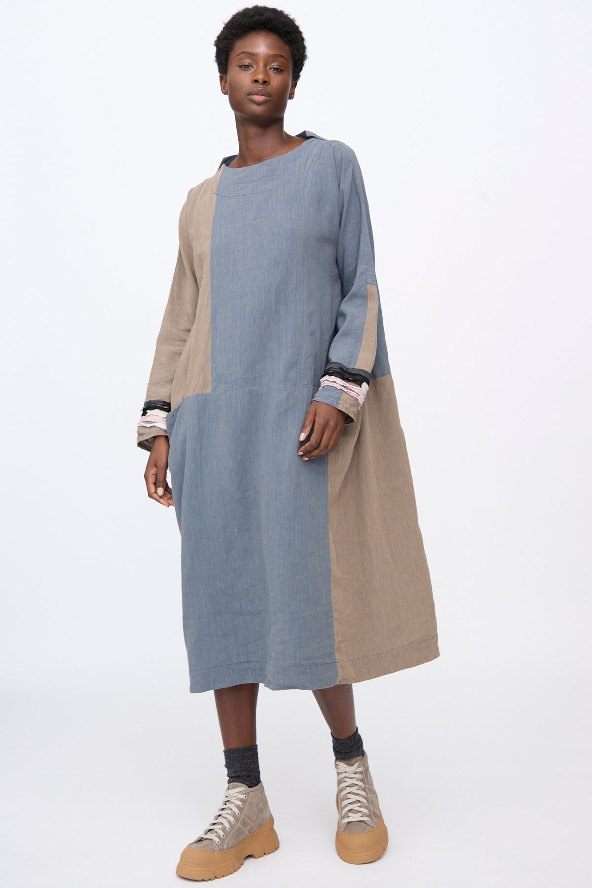A person wearing a Hallelujah Berger des Montagne de la Castille Dress in Blue Mix, a blue and beige color-blocked, linen blend robe with gathered cuffs and beige high-top sneakers.