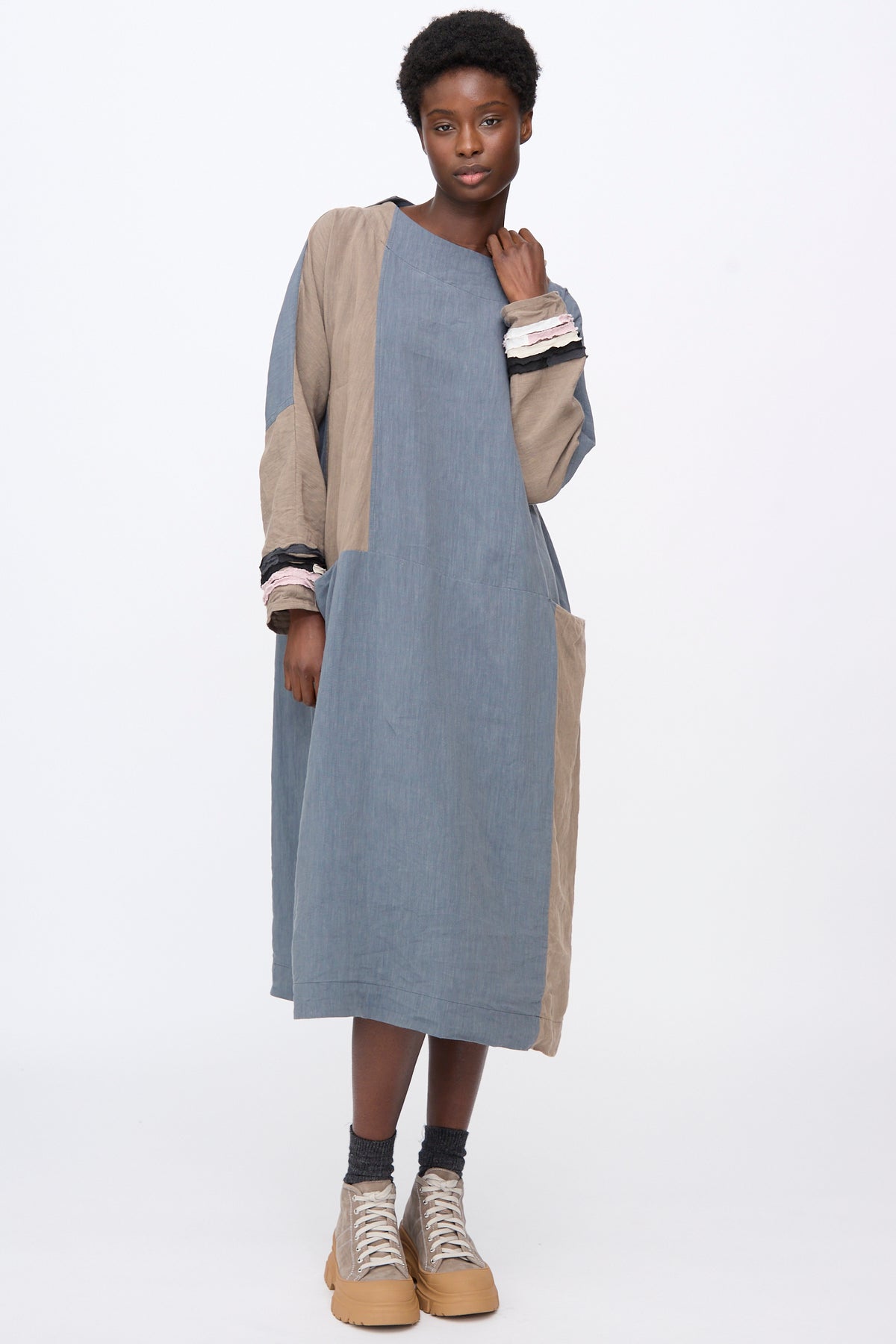 A person wears a Hallelujah Berger des Montagne de la Castille Dress in Blue Mix, a color-blocked midi linen blend robe with tiered ruffles at the cuffs, paired with platform sneakers.