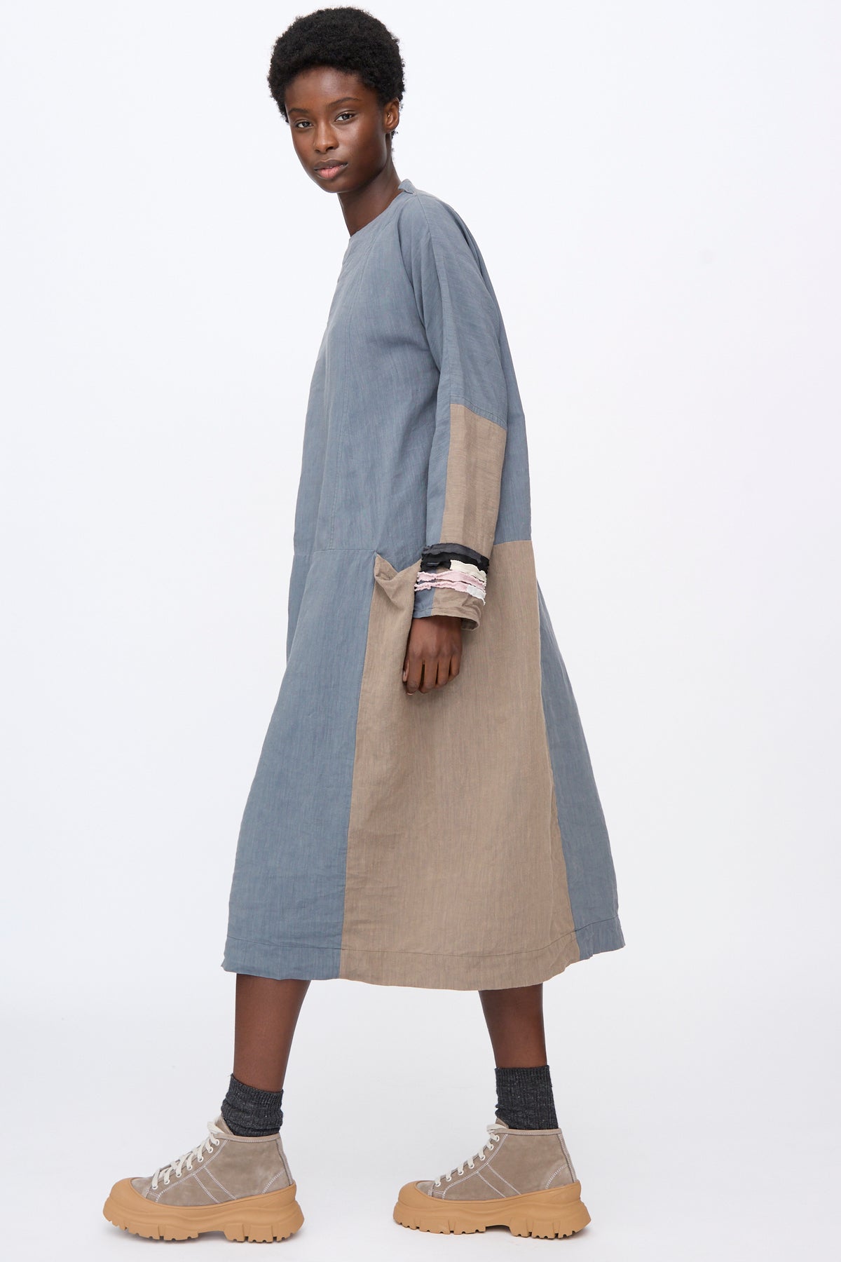 A person wears a blue and tan color-blocked Hallelujah Berger des Montagne de la Castille Dress in Blue Mix with long sleeves and tan high-top sneakers.