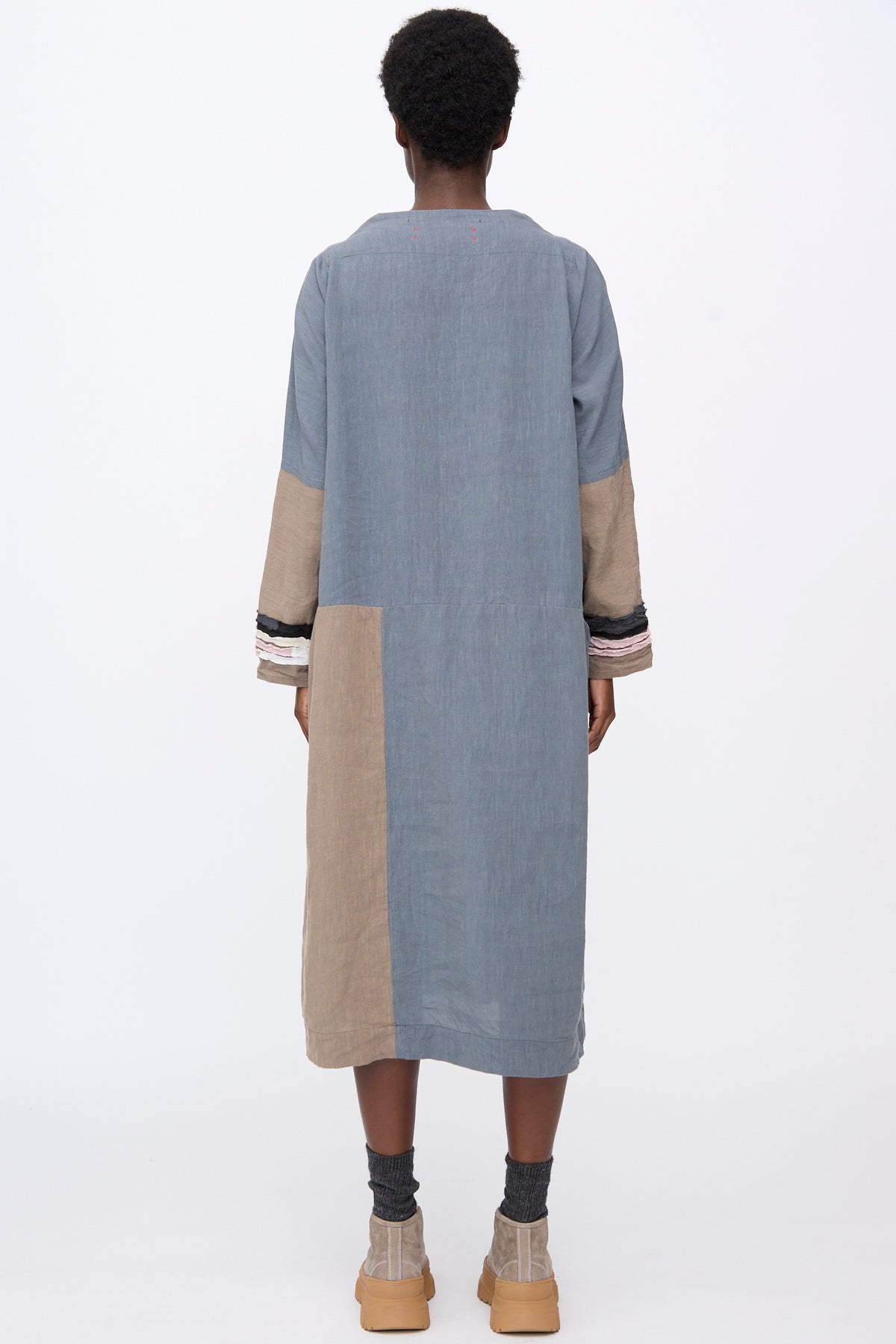Back view of a model wearing a knee-length, long-sleeved, color-blocked Hallelujah Berger des Montagne de la Castille Dress in Blue Mix with gathered cuffs, gray socks, and tan platform shoes.