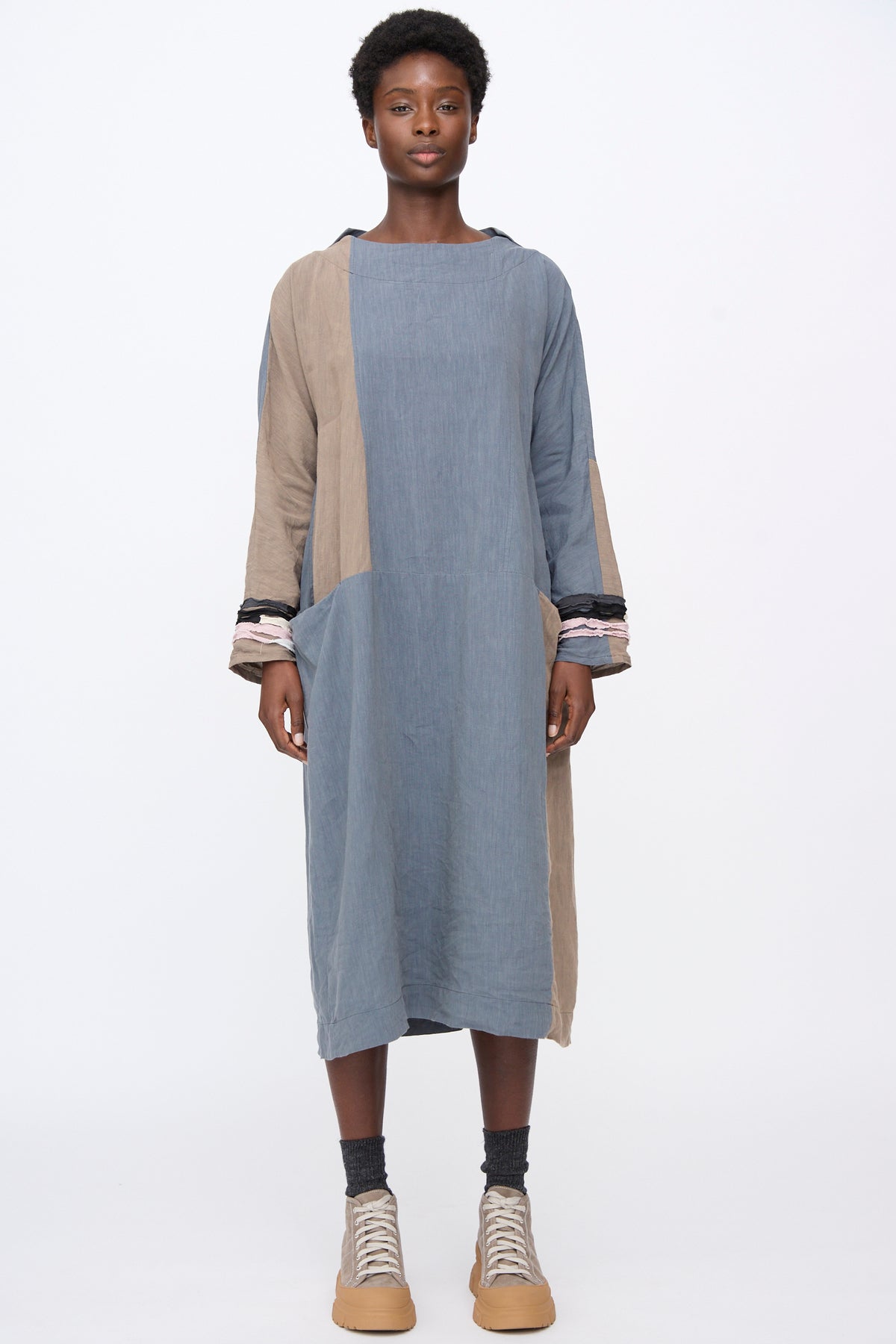A person wearing a Hallelujah Berger des Montagne de la Castille Dress in Blue Mix, a blue and tan color-blocked linen blend robe with knee-length, long-sleeved, ruffled cuffs.