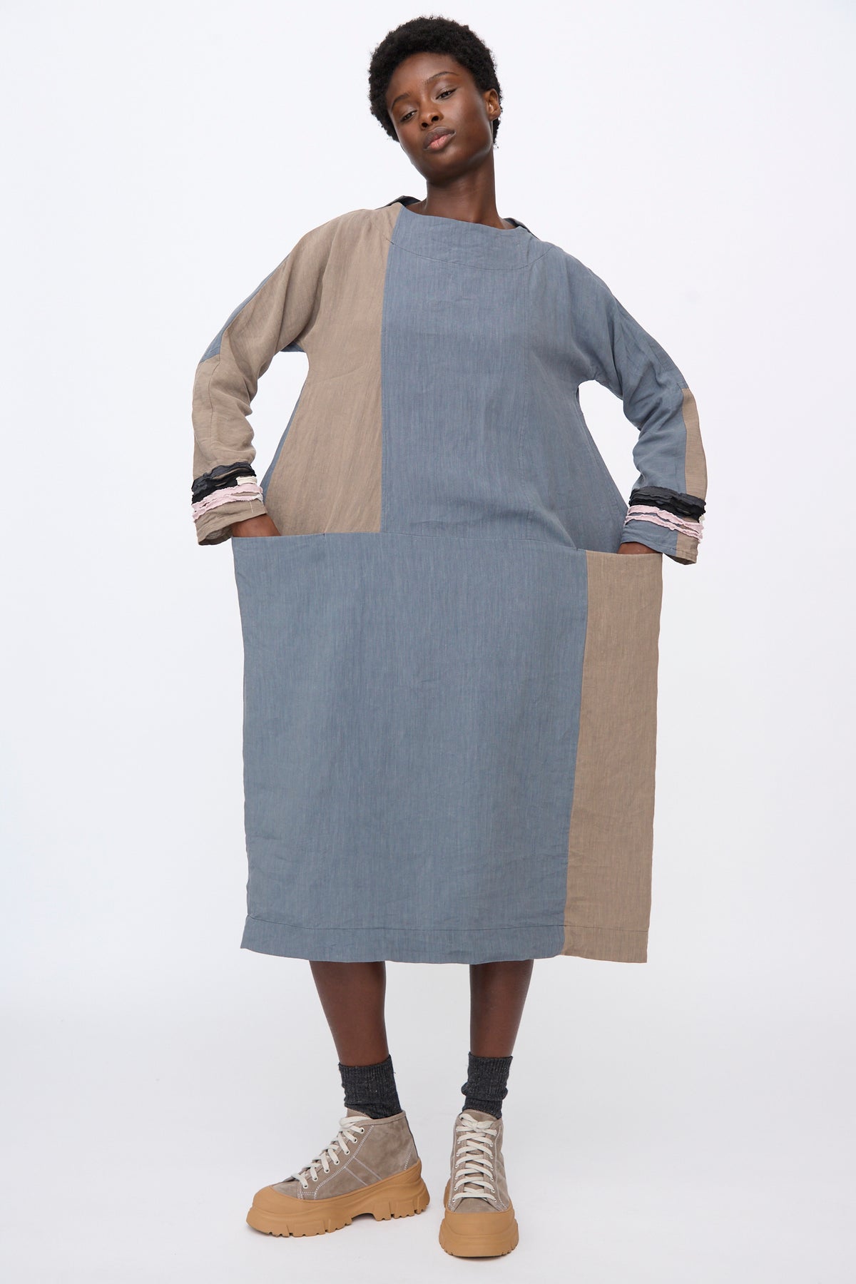 A person wears the Hallelujah Berger des Montagne de la Castille Dress in Blue Mix, a color-blocked linen blend midi dress in blue and tan with long sleeves, gathered cuffs, and side pockets.