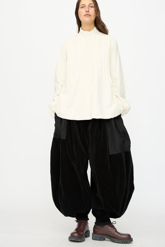 A person stands against a plain background wearing a loose white button-up shirt and Hallelujah's Bloomer in Black—oversized velveteen cotton pants with balloon legs—paired with brown lace-up boots.