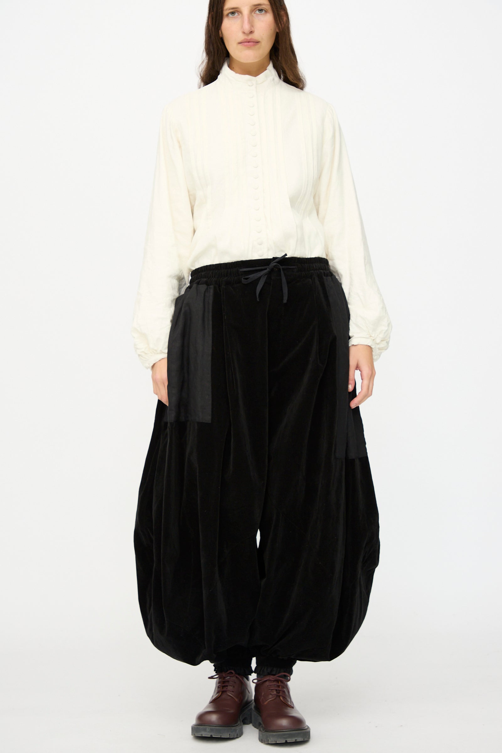 A person stands against a plain background wearing Hallelujah's Bloomer in Black, paired with a high-neck white blouse and brown lace-up boots.