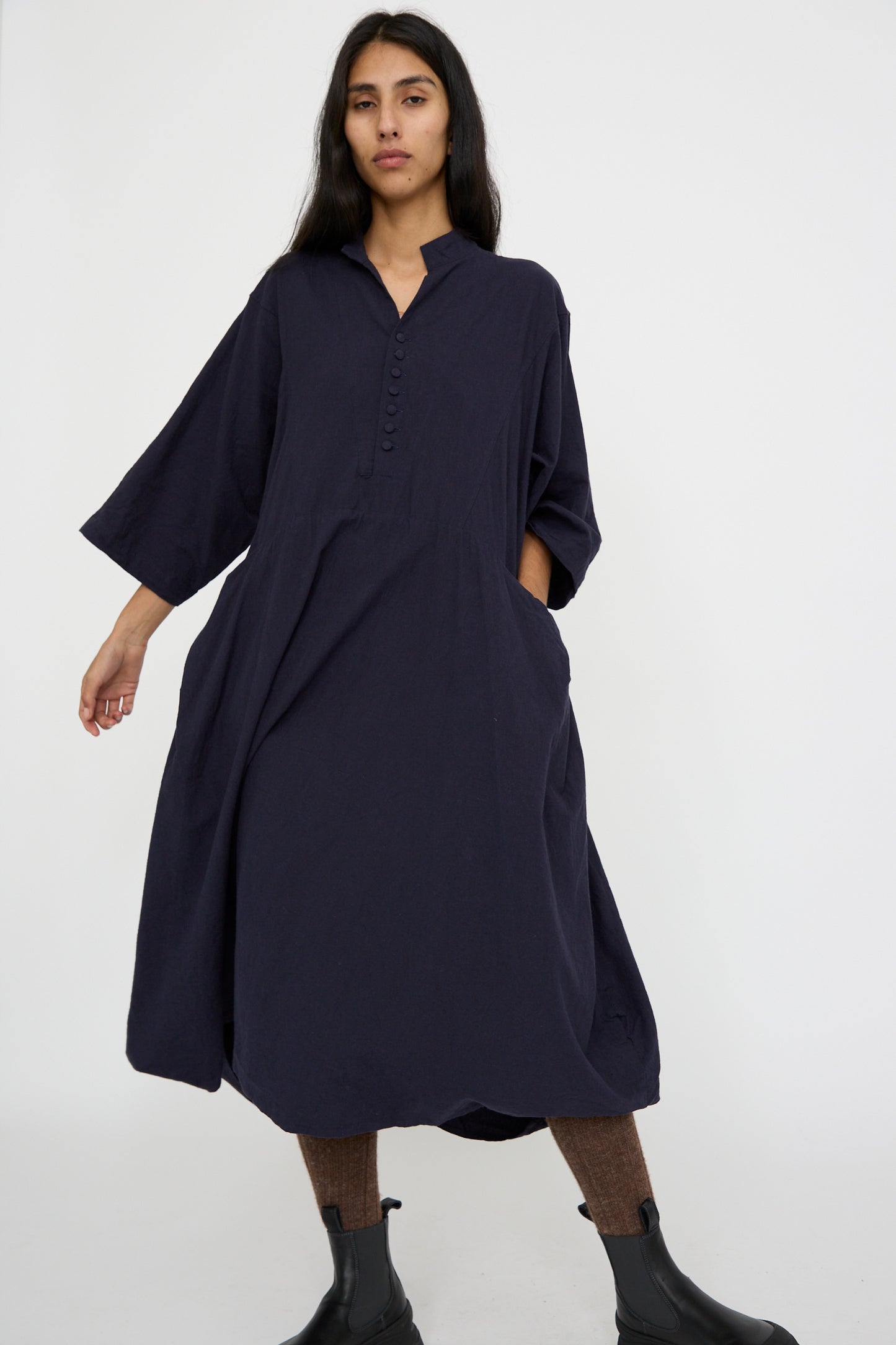 A person stands against a plain background, wearing a relaxed-fit Canvas Acolyte Robe in Navy by Hallelujah, featuring a buttoned neckline and subtle bell sleeves, crafted from a linen wool blend.