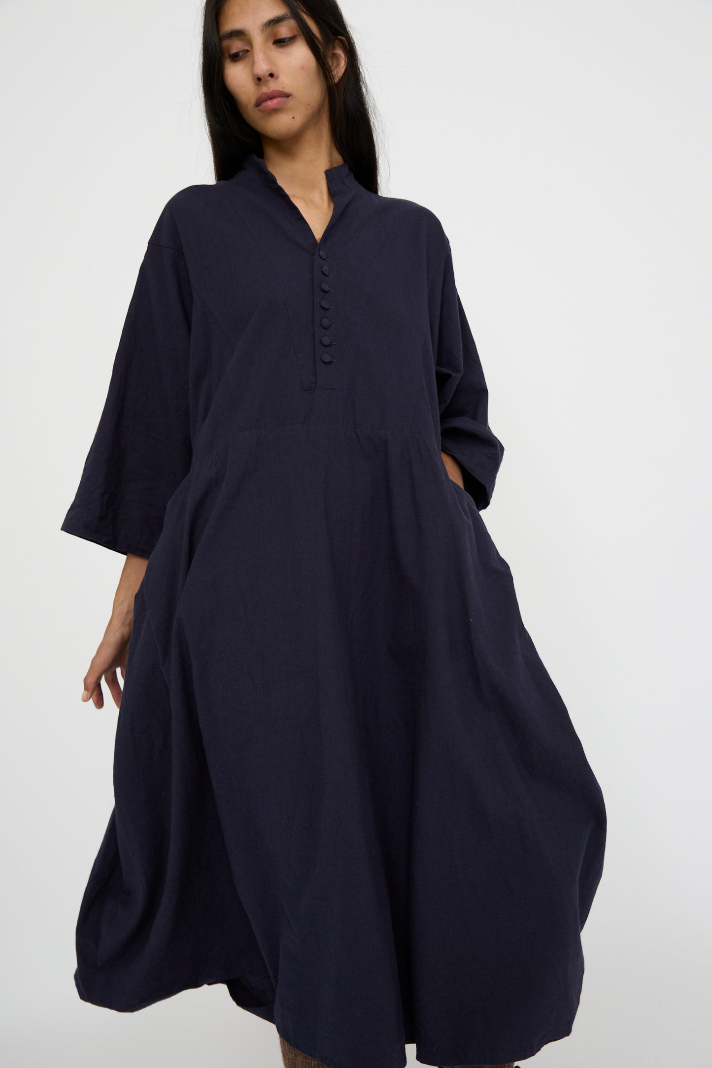 A person is wearing the Canvas Acolyte Robe in Navy by Hallelujah, a relaxed-fit dress crafted from a linen wool blend with three-quarter bell sleeves and front buttons, standing against a plain background.