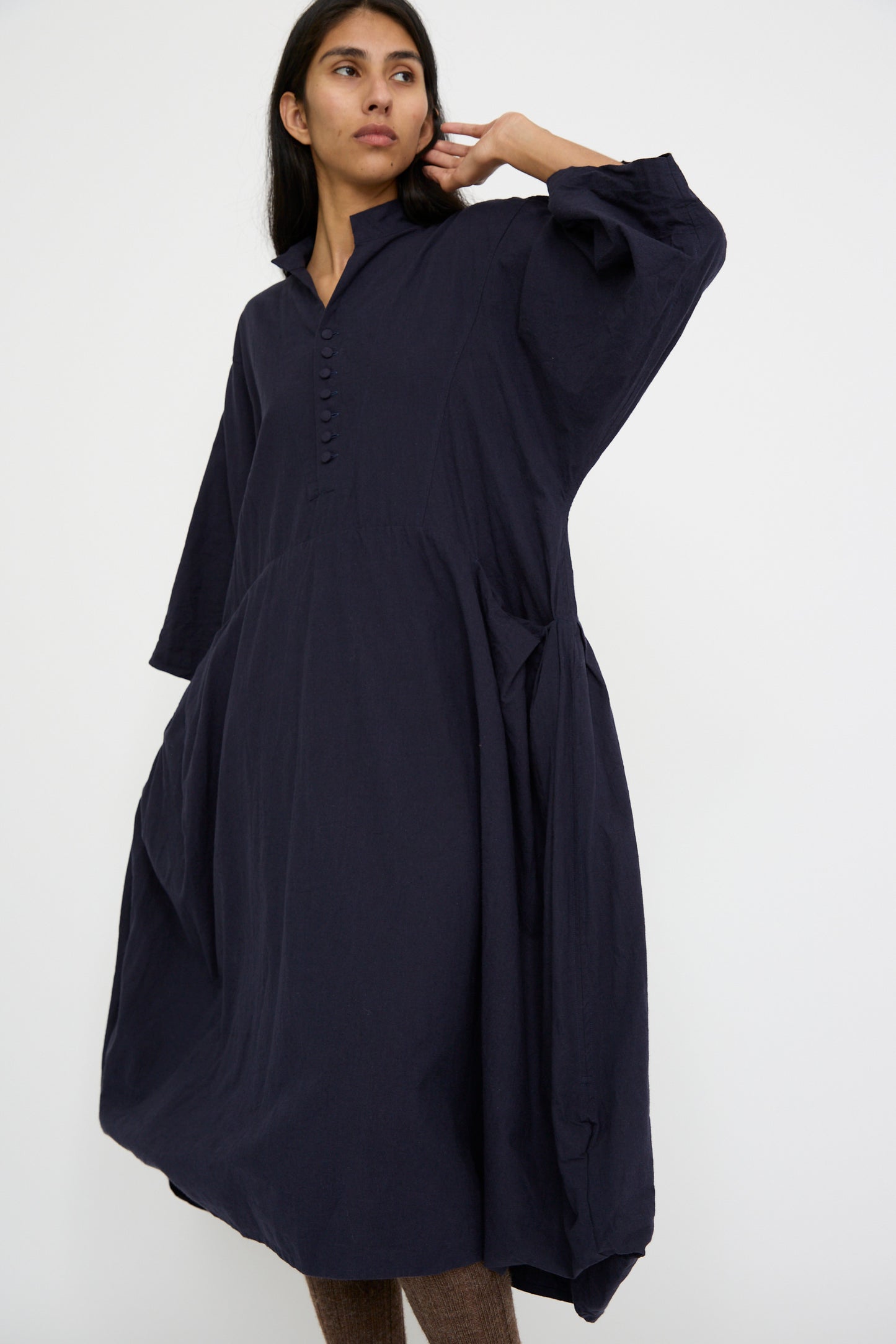 A person wearing a relaxed fit Canvas Acolyte Robe in Navy made from a linen wool blend, designed by Hallelujah and featuring bell sleeves, stands against a plain background. They have long hair and one hand near their neck.