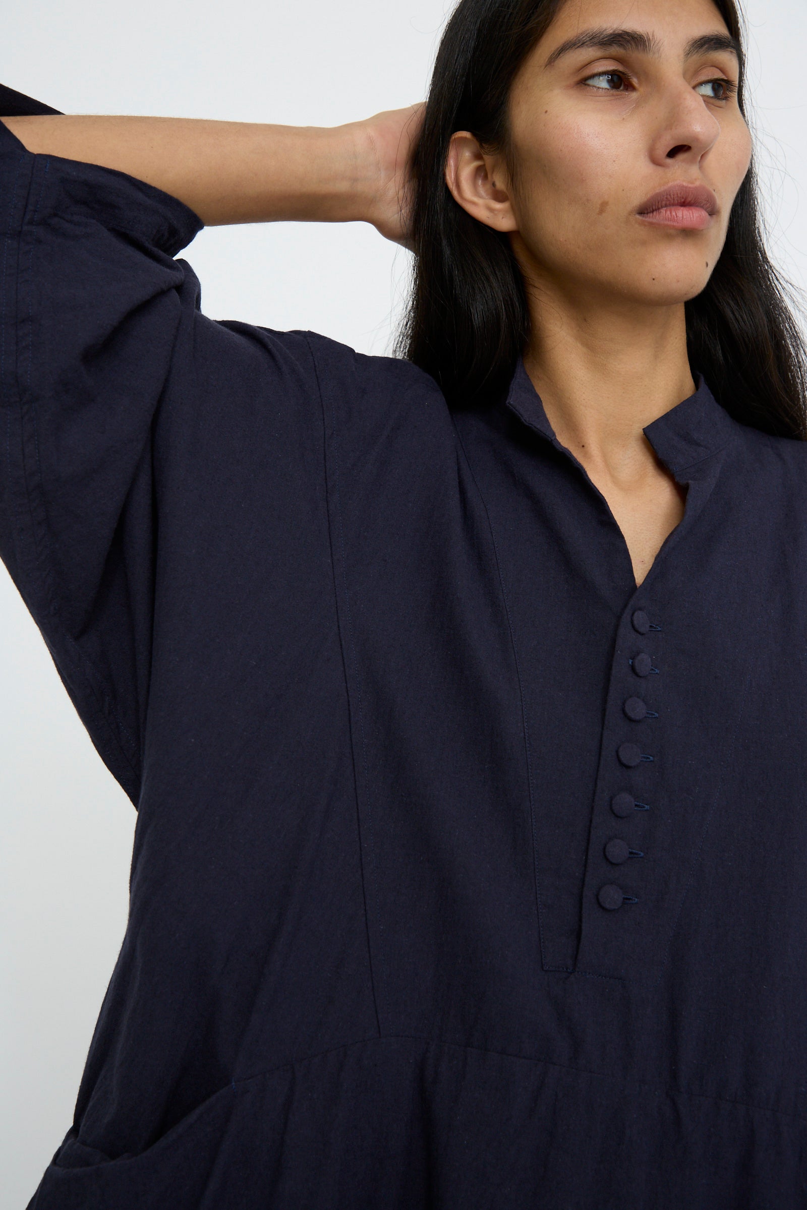 A person with long hair wearing the Hallelujah Canvas Acolyte Robe in Navy, which has buttoned details and bell sleeves, gazes to the side with one arm raised.