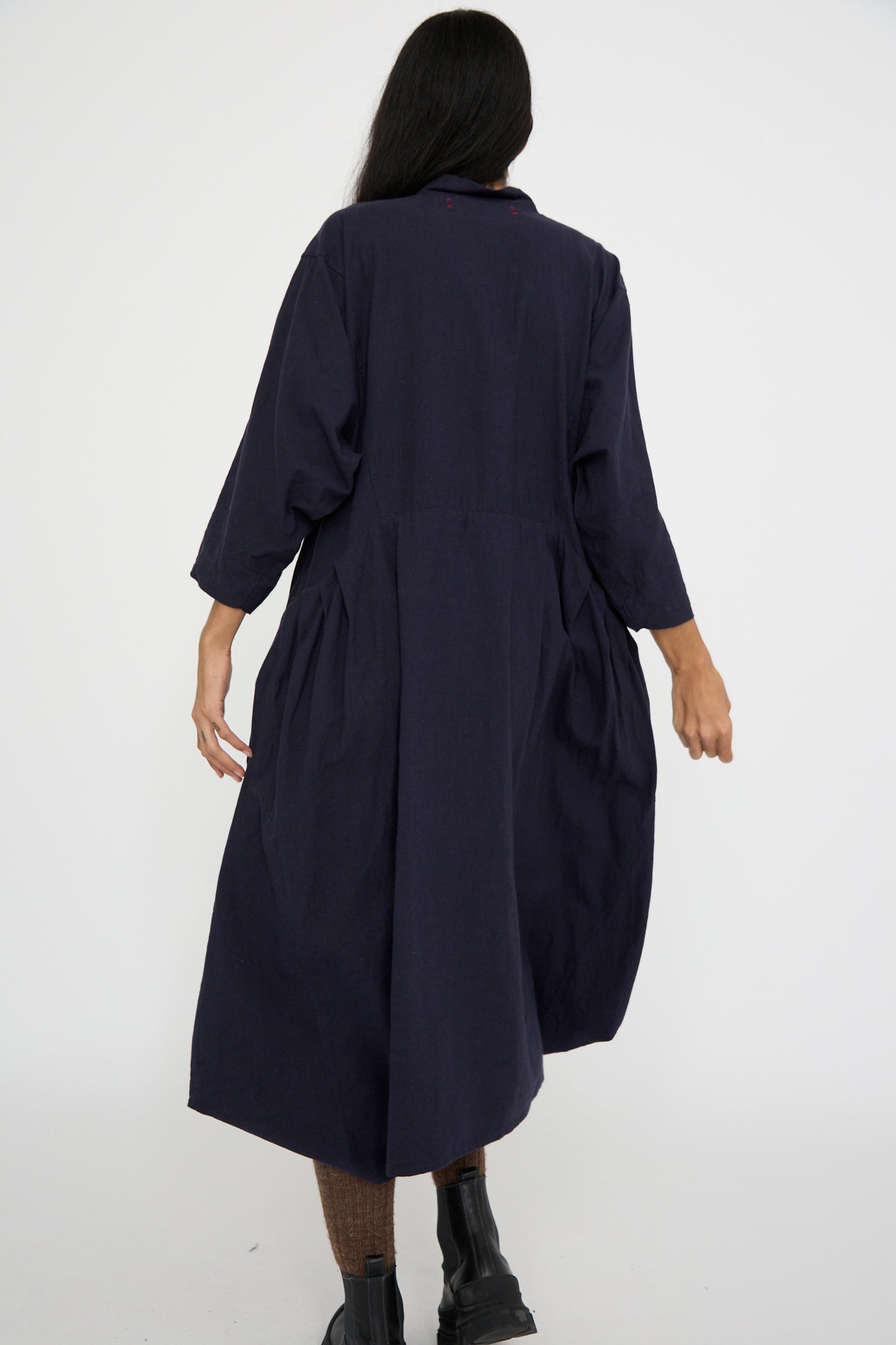 A person with long dark hair is seen from behind, dressed in the Canvas Acolyte Robe in Navy by Hallelujah, paired with brown pants and black boots, against a plain white background.