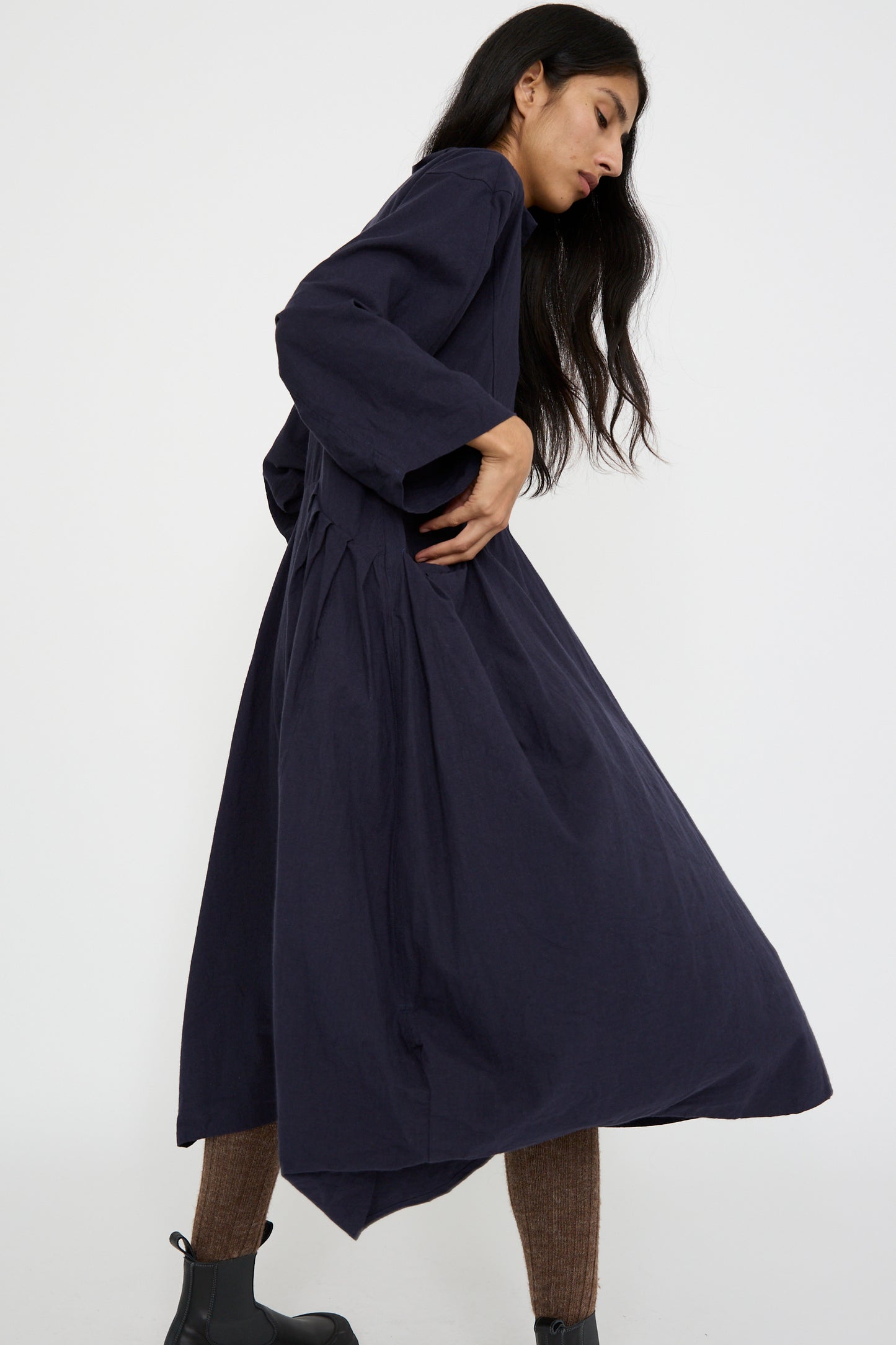 A person wearing the Canvas Acolyte Robe in Navy by Hallelujah, featuring a long, dark linen wool blend with relaxed fit and bell sleeves, stands in profile against a plain background, paired with brown tights.