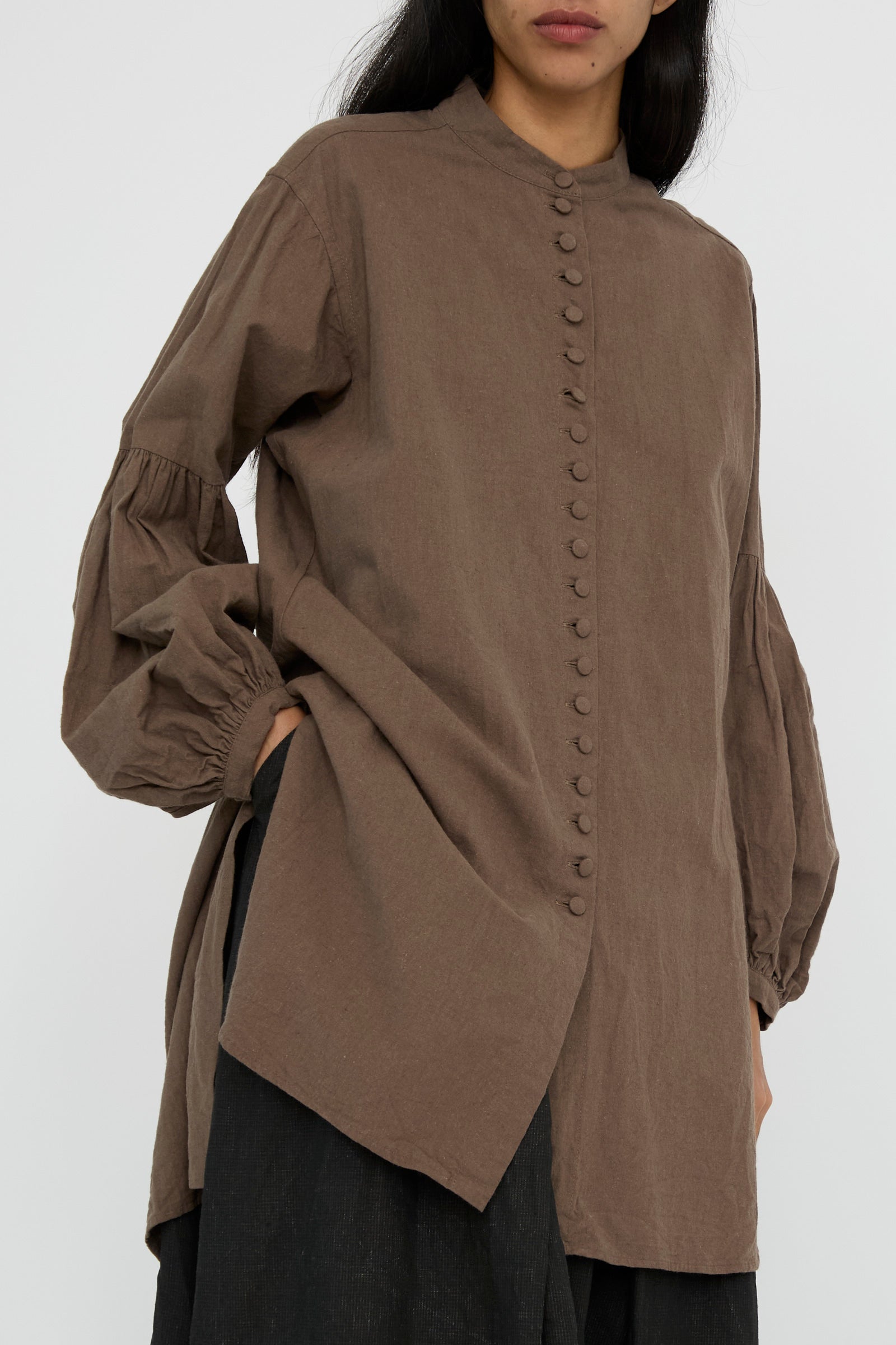 An individual is dressed in a Canvas Chemise Religieuse in Chocolate by Hallelujah, featuring bishop sleeves, paired with dark pants against a plain background.