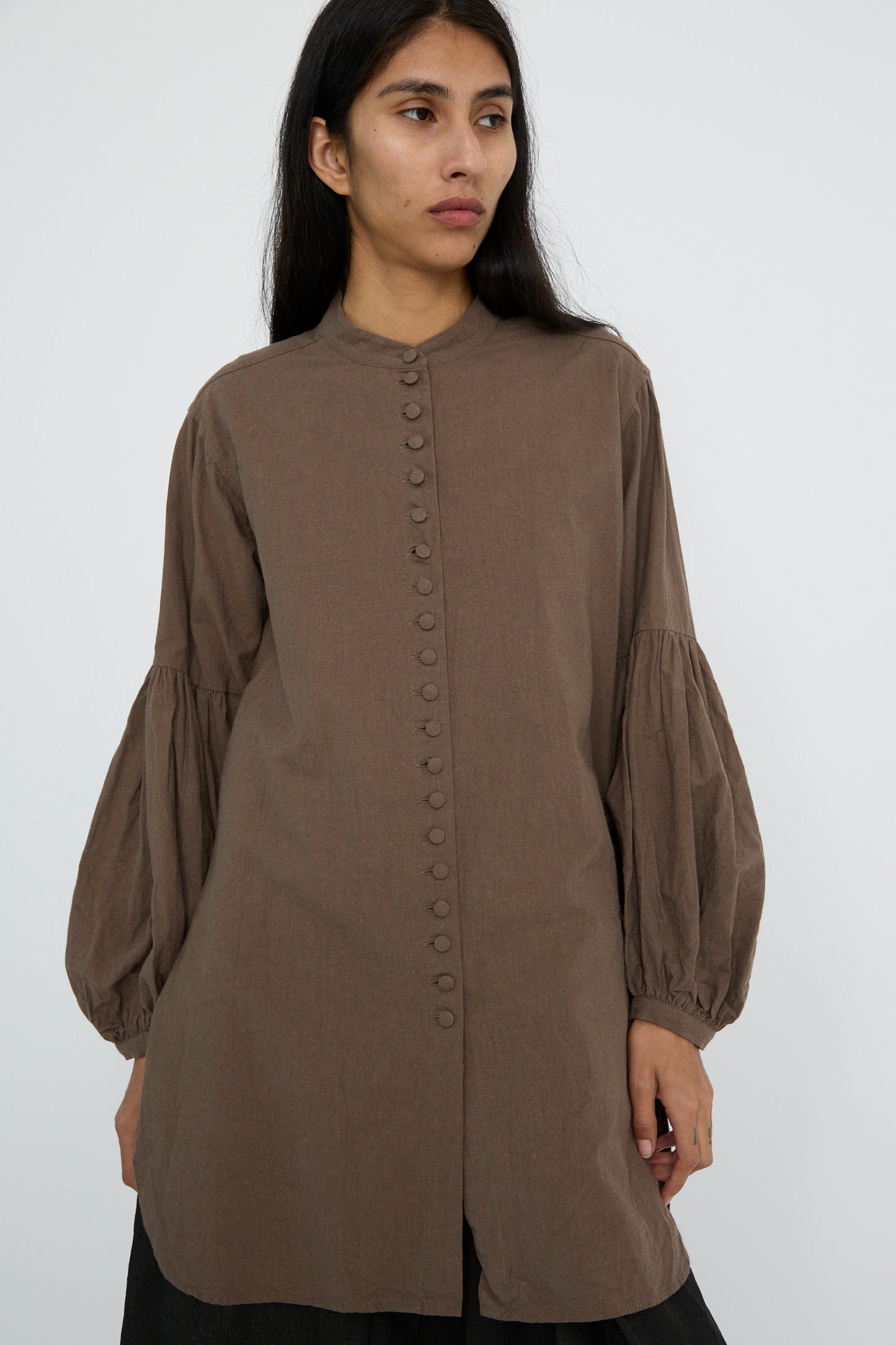 A person is wearing the Canvas Chemise Religieuse in Chocolate with bishop sleeves from Hallelujah, featuring a band collar and puffed sleeves, and looking to the side against a plain white background.