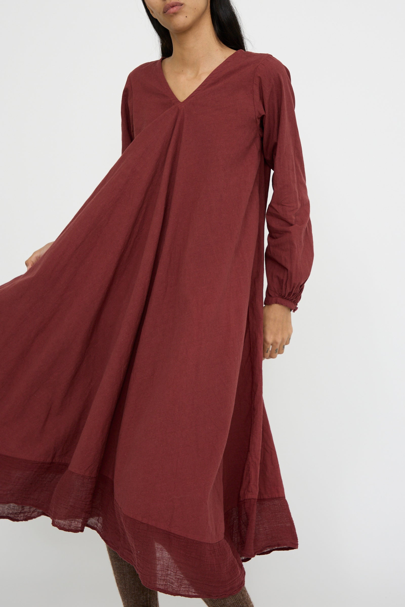 A person is wearing the Canvas Robe de Femme de Chambre by Hallelujah in Bordeaux, a long-sleeved maxi dress with a V-neck and flowing skirt against a white background.