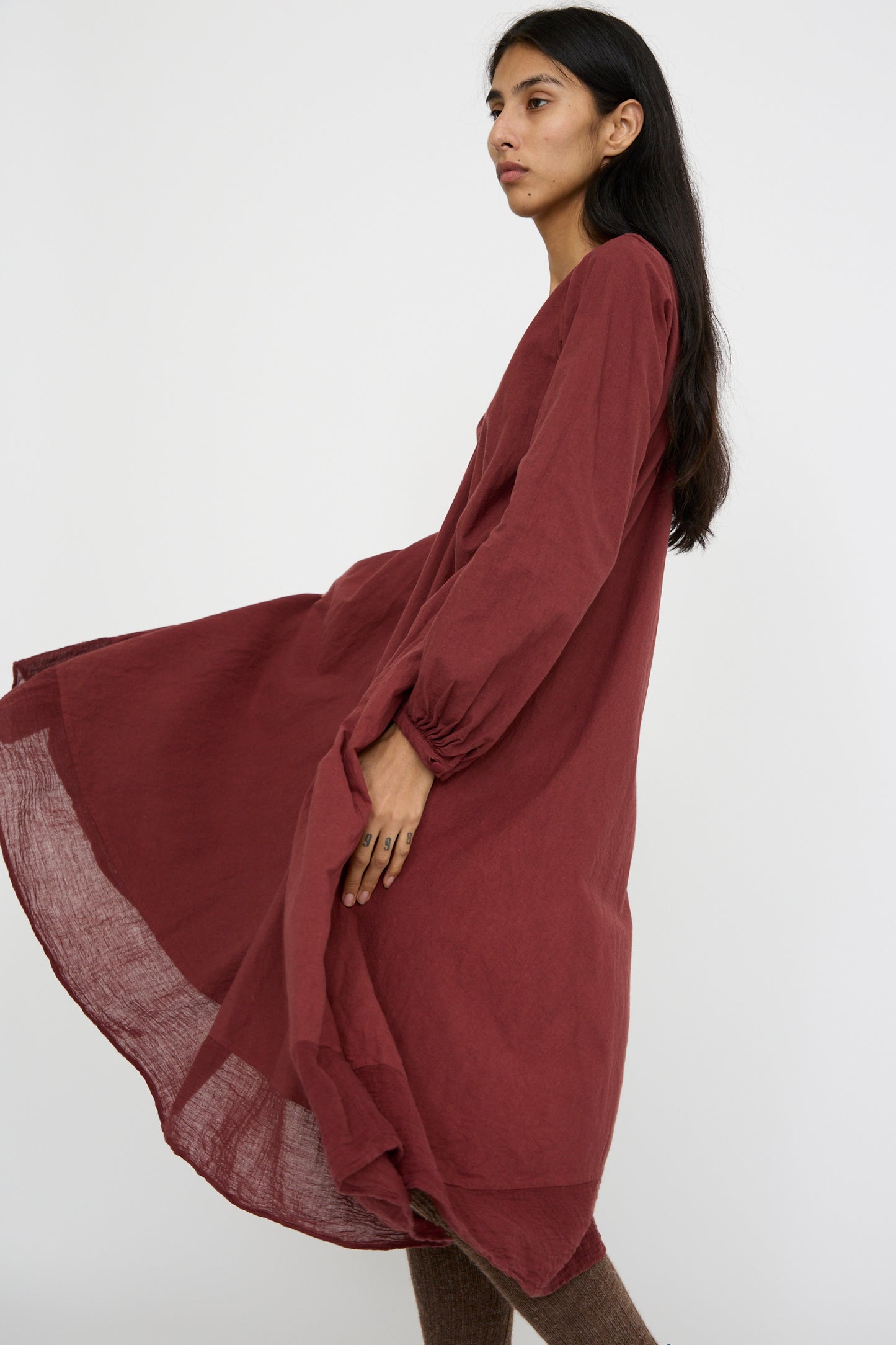 A person with long dark hair wearing Hallelujah's Canvas Robe de Femme de Chambre in Bordeaux, showcasing peasant sleeves, stands against a plain background.