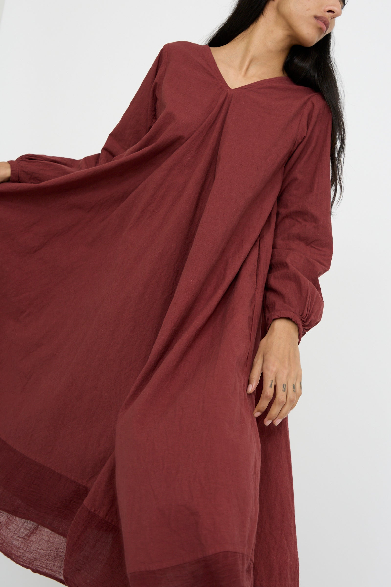 A person wearing Hallelujah's Canvas Robe de Femme de Chambre in Bordeaux, featuring peasant sleeves, stands against a plain backdrop, holding the flowing dress out with one hand.