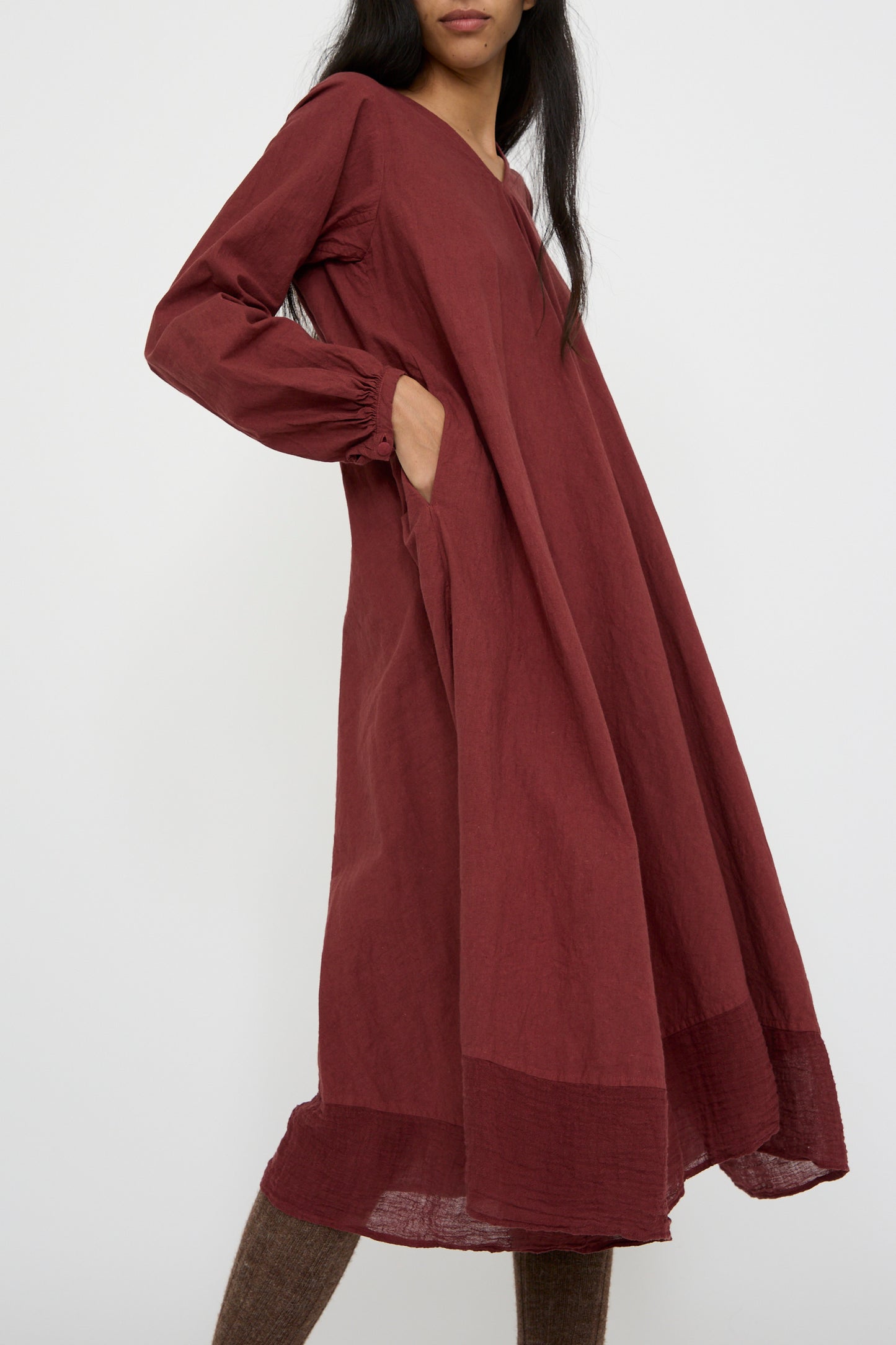 A person is wearing the Canvas Robe de Femme de Chambre in Bordeaux by Hallelujah, which is a long, loose maxi dress featuring peasant sleeves and pockets, standing against a plain background with a hand in a pocket. 