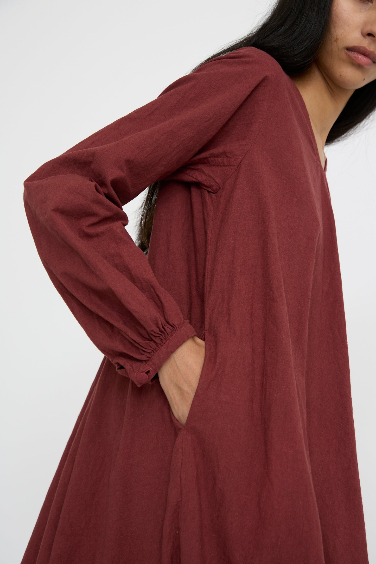 A person models the Hallelujah Canvas Robe de Femme de Chambre in Bordeaux, featuring peasant sleeves and a comfortable pocket, set against a plain backdrop.