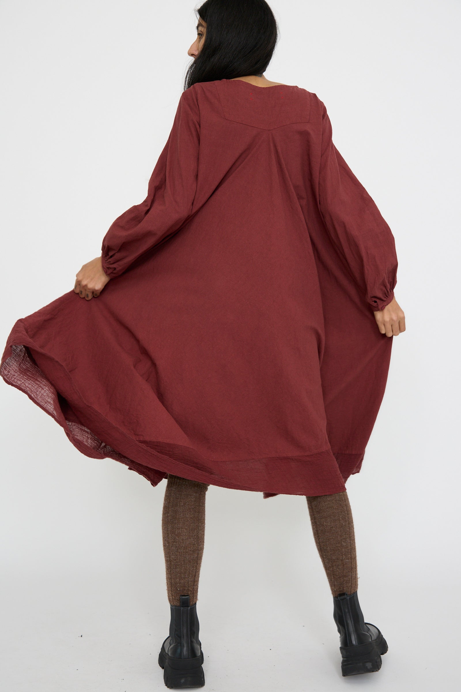 A person wearing the Hallelujah Canvas Robe de Femme de Chambre in Bordeaux, complemented by brown leggings and black boots, stands facing away against a plain background.