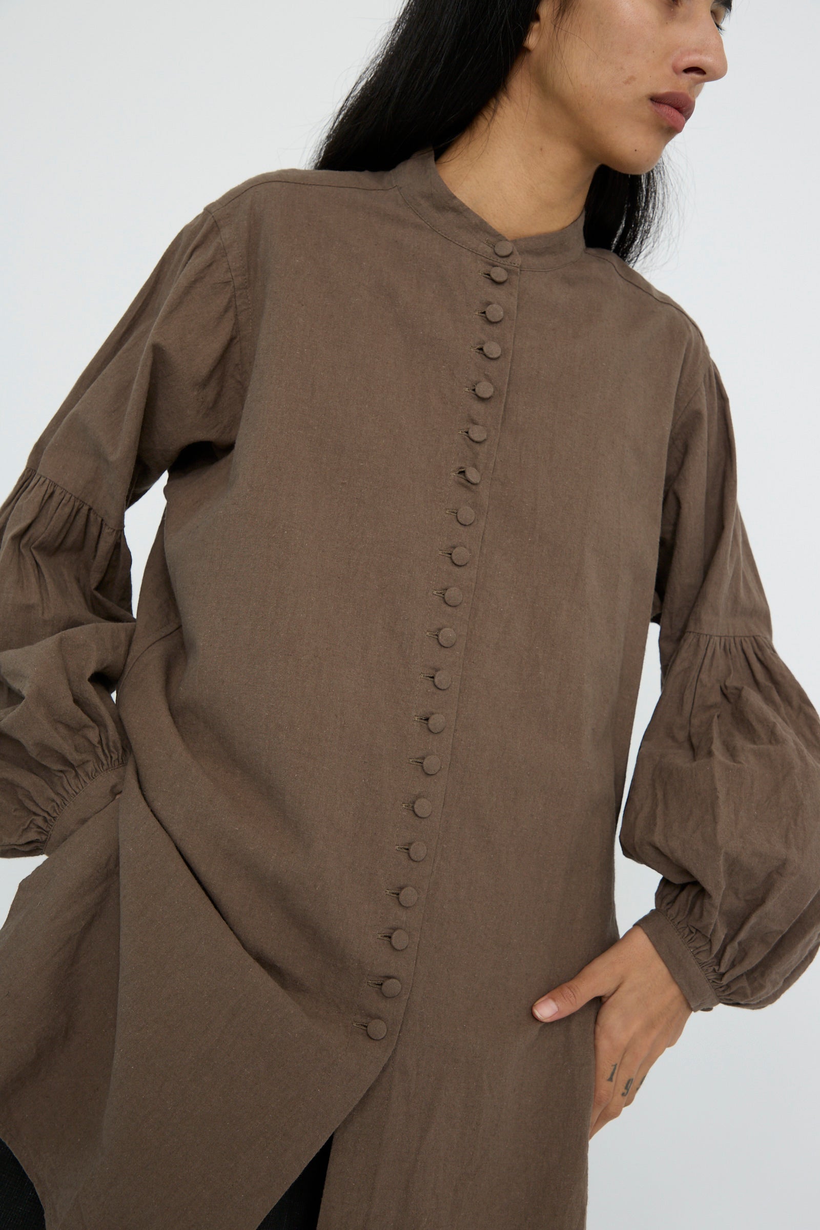 A person wearing the Canvas Chemise Religieuse in Chocolate by Hallelujah, featuring a band collar and bishop sleeves, stands against a plain background.