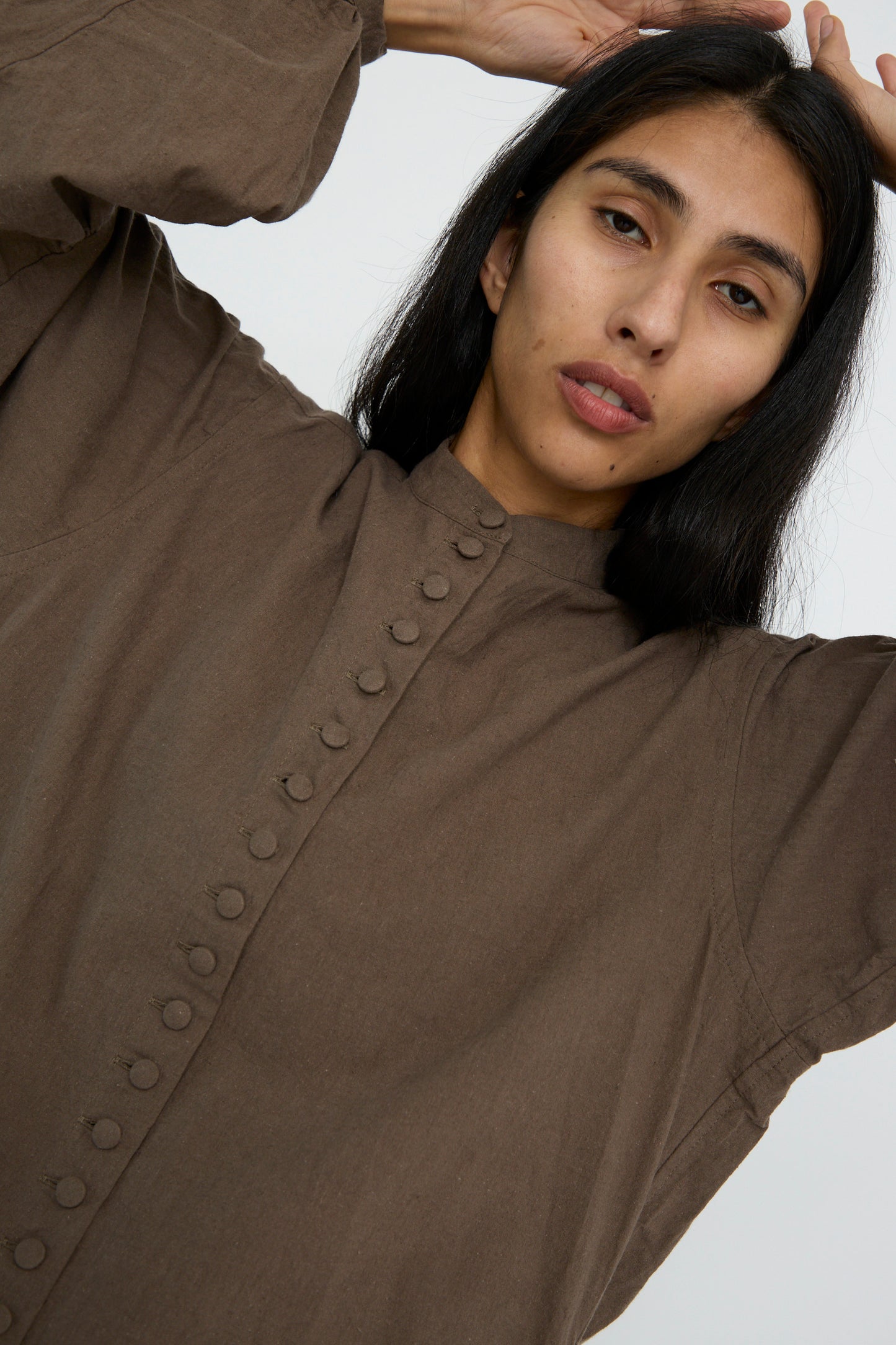 A person with long, dark hair poses with hands above their head against a plain background, wearing the Hallelujah Canvas Chemise Religieuse in Chocolate.