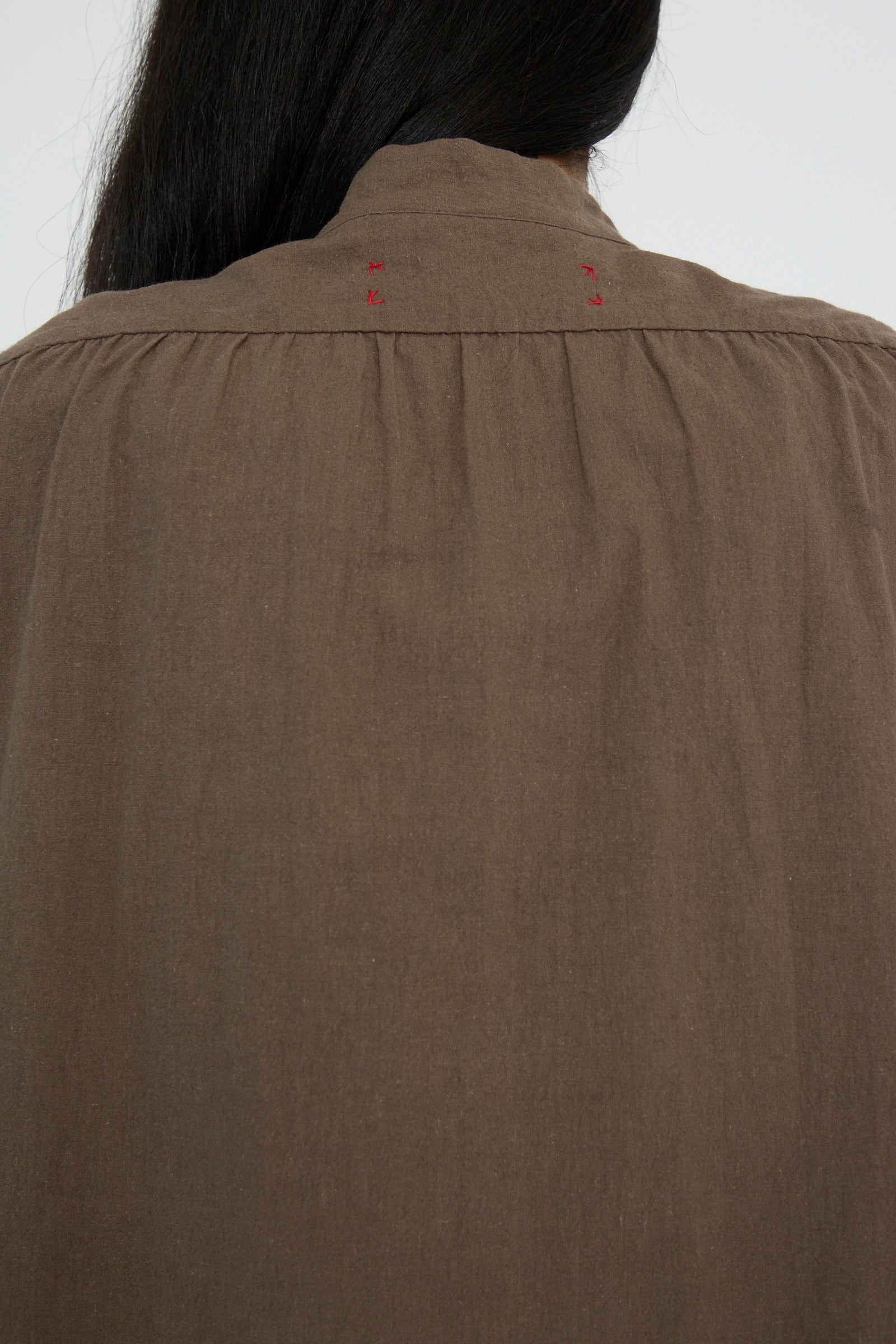 A close-up back view of a person with long dark hair is wearing the Canvas Chemise Religieuse in Chocolate by Hallelujah, featuring small red embroidery near the band collar.