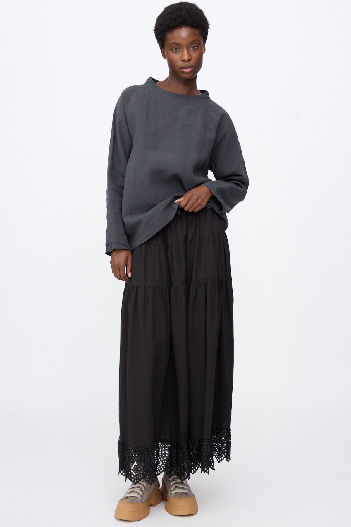 A model wears a gray Chemise de Berger in Gray long-sleeved linen blend shirt and a long black tiered skirt with a lace hem, paired with tan sneakers.