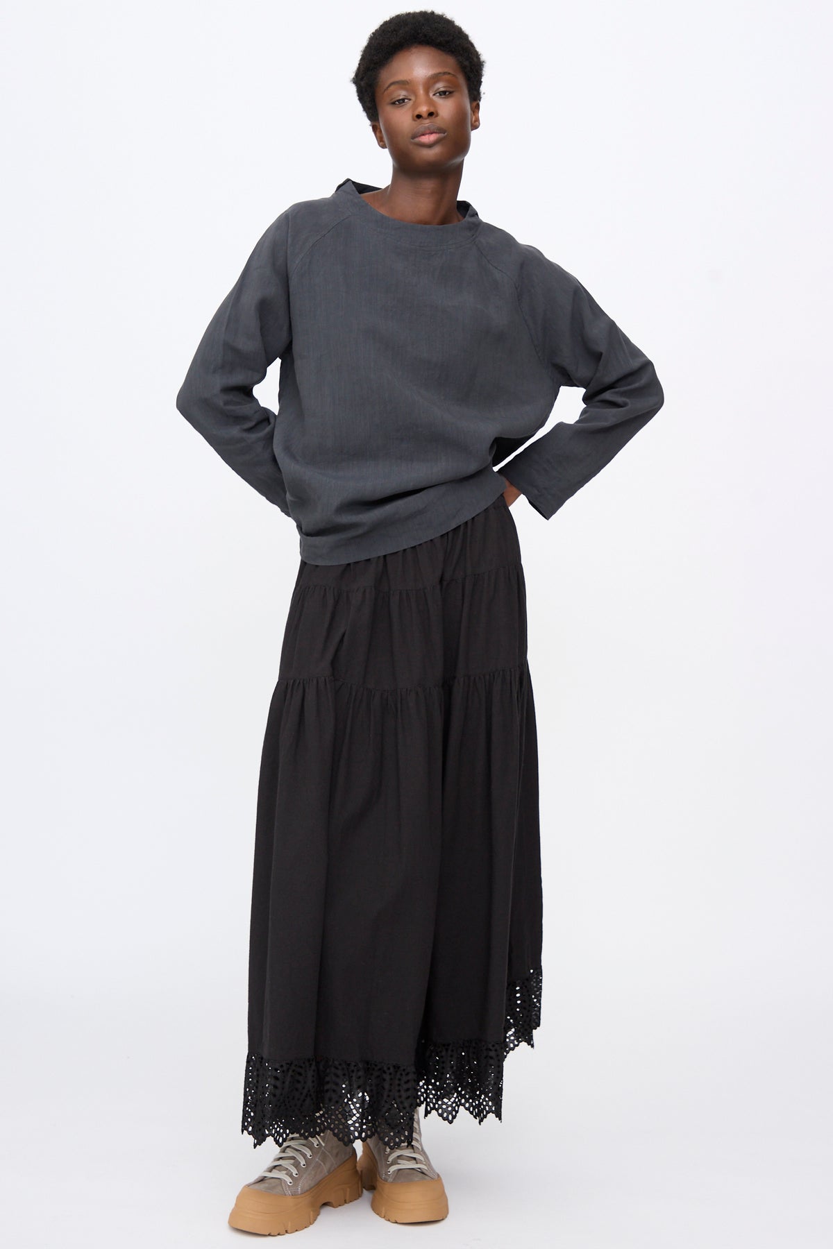A person wearing a charcoal gray Hallelujah Chemise de Berger in Gray, a black tiered maxi skirt with a lace hem, and beige platform sneakers.
