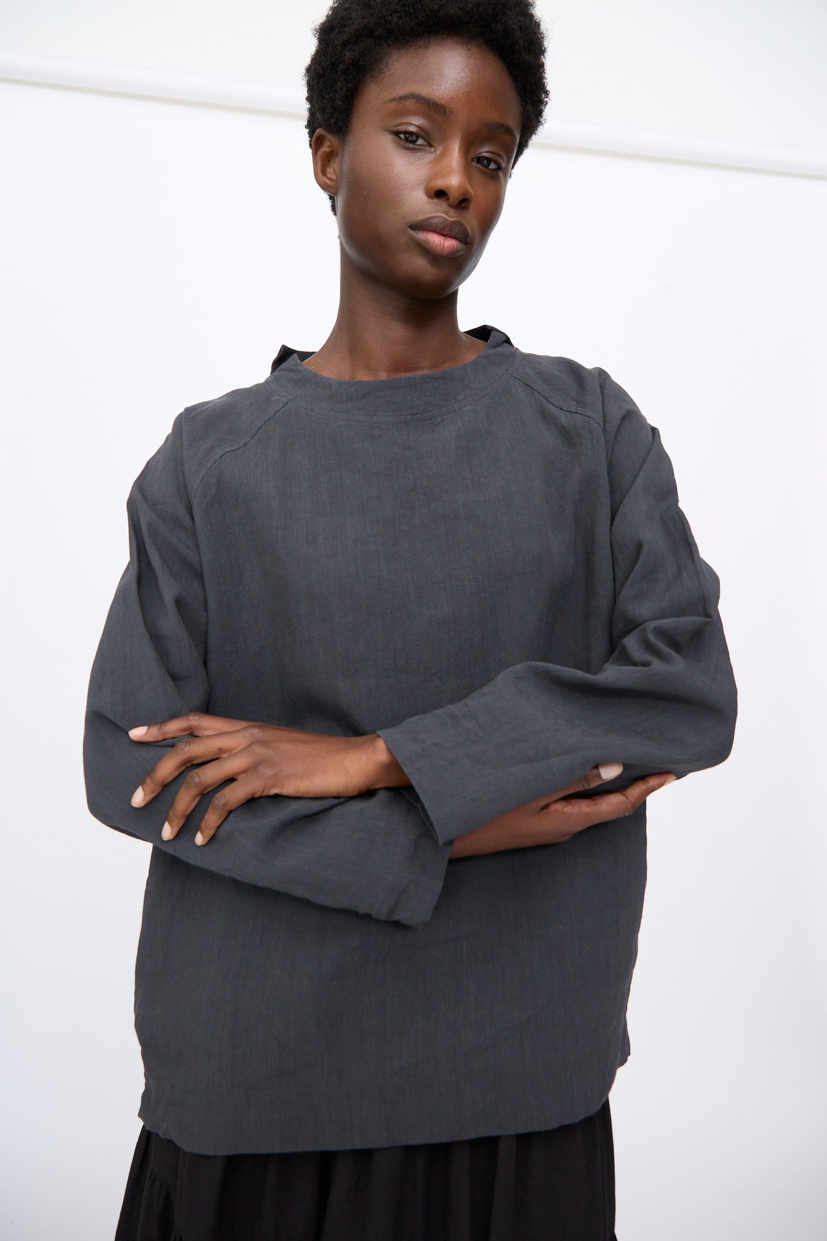 A person wearing a Chemise de Berger in Gray, a long-sleeved gray linen blend shirt with a round neckline, stands with their arms crossed.