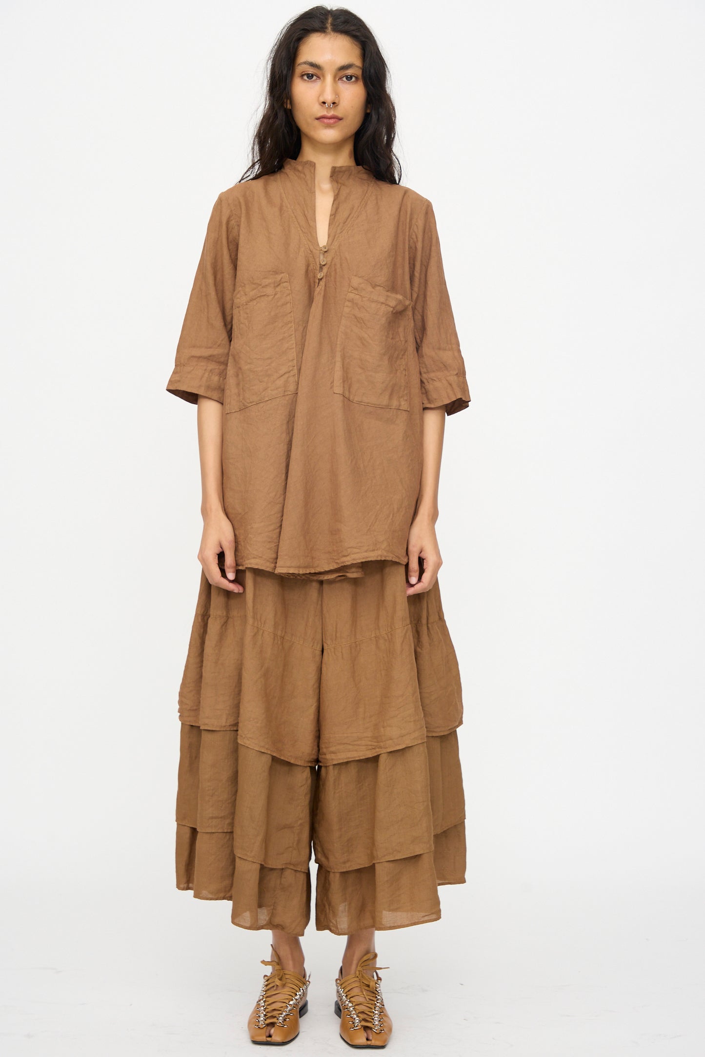 A person stands facing forward, wearing the Hallelujah Chemise de Garcon Berger in Persimmon Dye—a loose-fitting short sleeve blouse with large pockets—paired with layered brown wide-leg pants and woven shoes.
