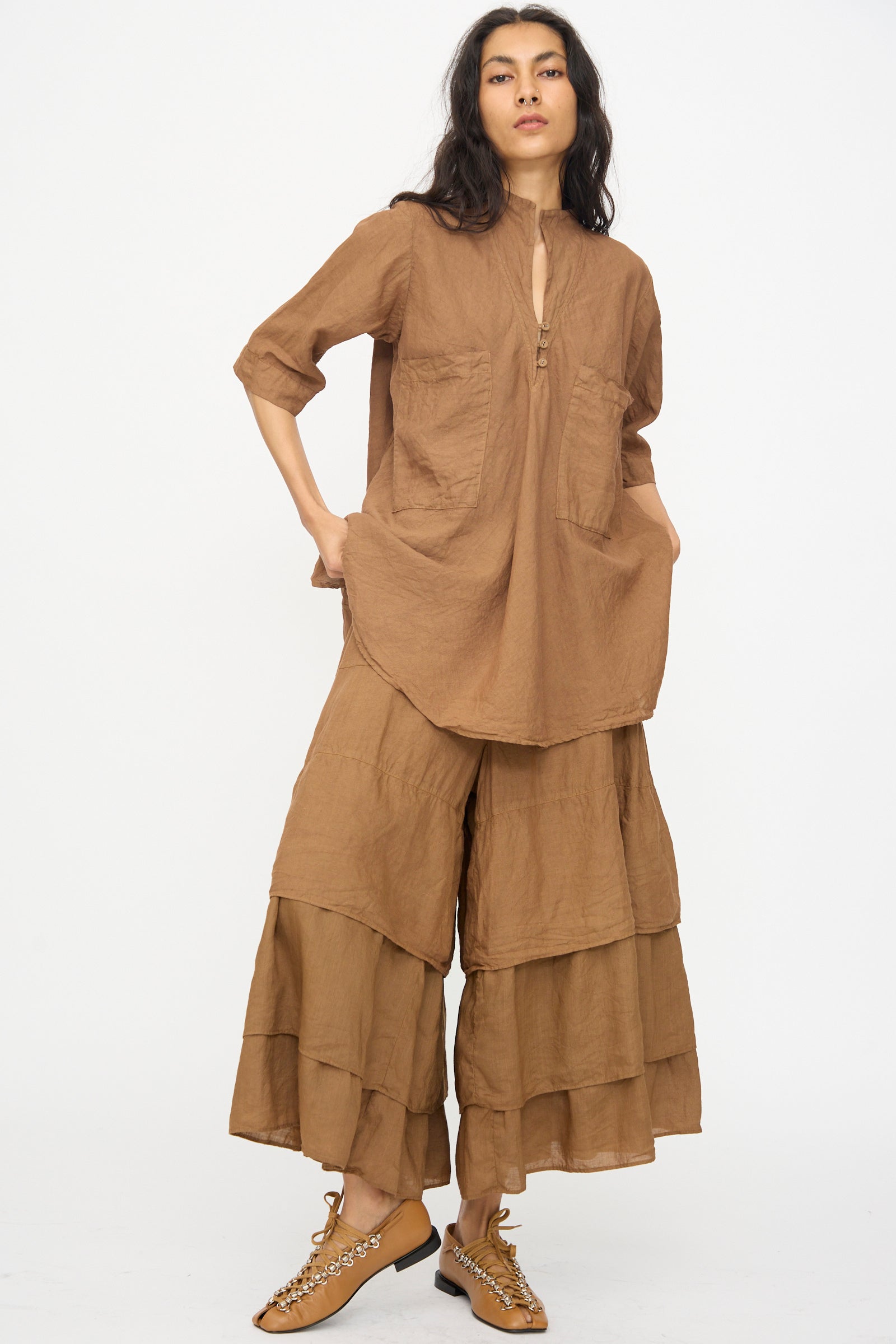 A person stands against a white background, wearing the Hallelujah Chemise de Garcon Berger in Persimmon Dye with matching wide-legged, tiered trousers and tan lace-up shoes.