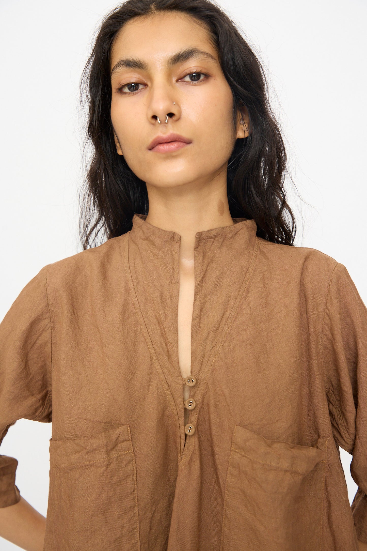 A person with long dark hair wears the Hallelujah Chemise de Garcon Berger in Persimmon Dye—a brown, short-sleeve blouse with front pockets—facing the camera against a plain background, blending elegance and comfort in a refined style.