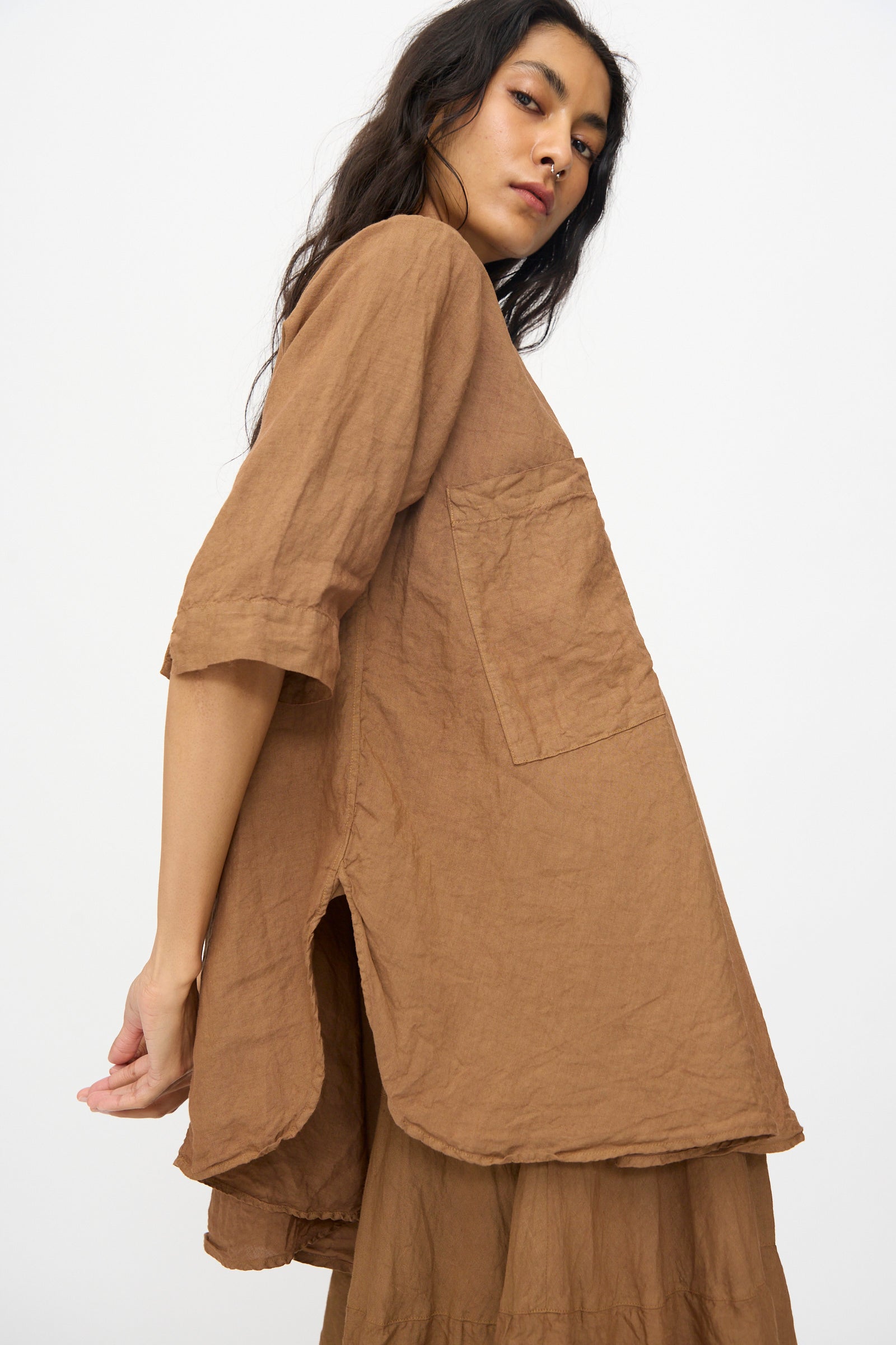 Model wears the Chemise de Garcon Berger in Persimmon Dye by Hallelujah—an easy, brown linen chambray blouse with a large front pocket and side slit—posed against a plain white background.