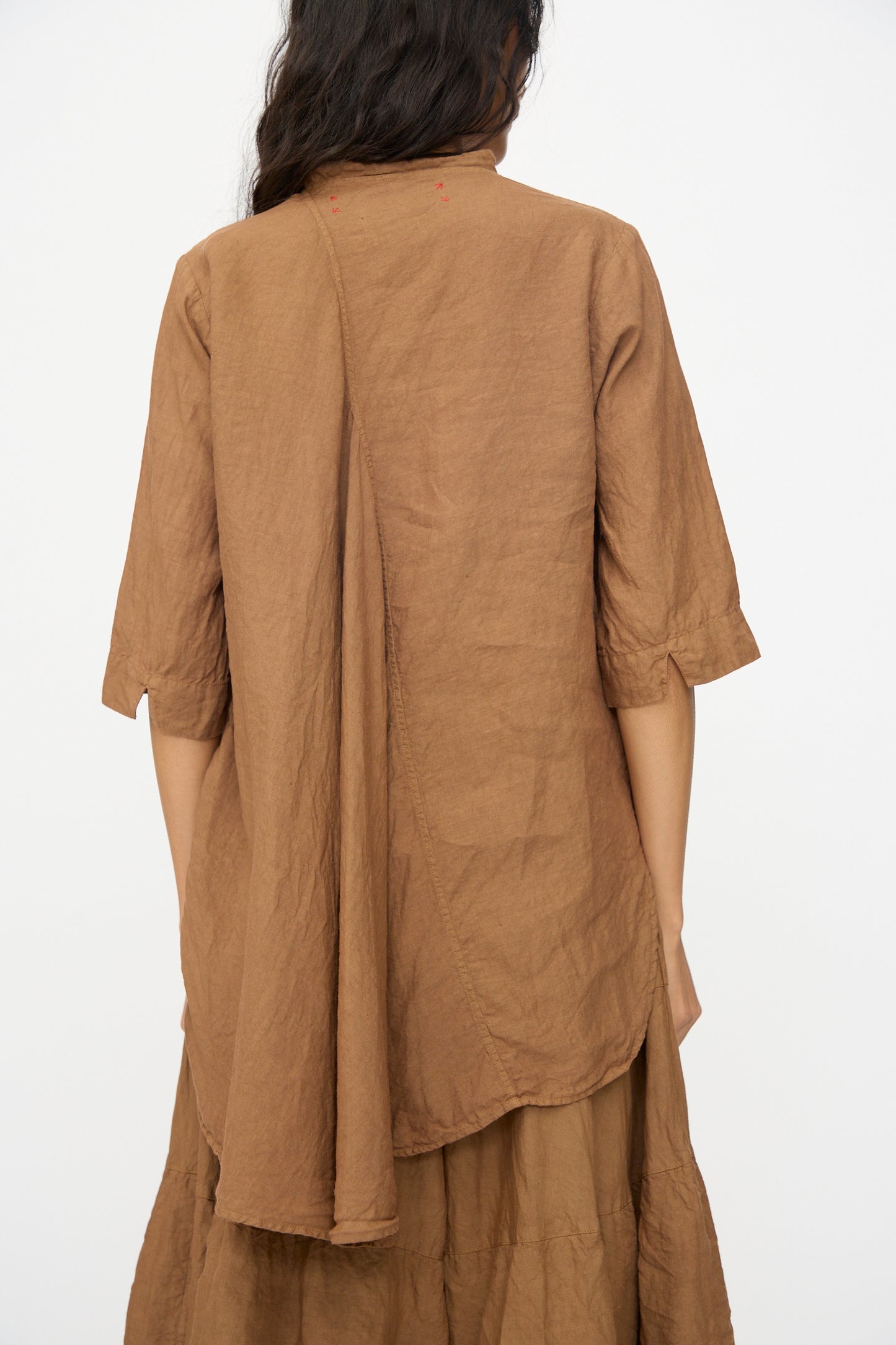 A person with long dark hair, facing away, wears the Hallelujah Chemise de Garcon Berger in persimmon dye—a loose-fitting brown linen blouse with asymmetric back seam and matching skirt, made in Japan.