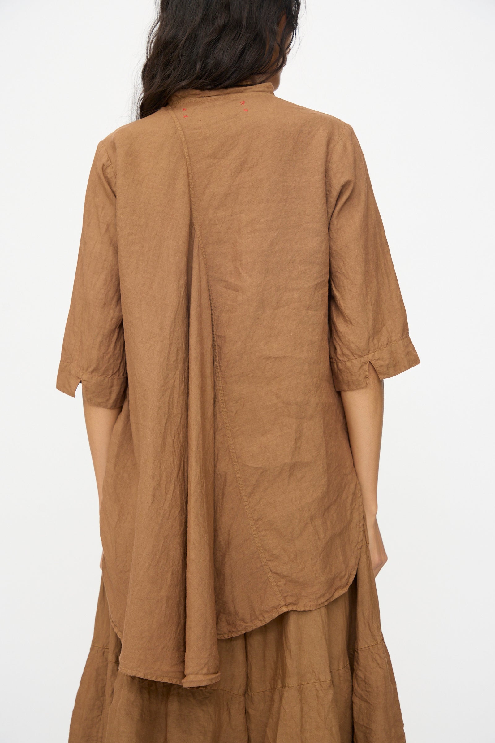 A person with long dark hair, facing away, wears the Hallelujah Chemise de Garcon Berger in persimmon dye—a loose-fitting brown linen blouse with asymmetric back seam and matching skirt, made in Japan.