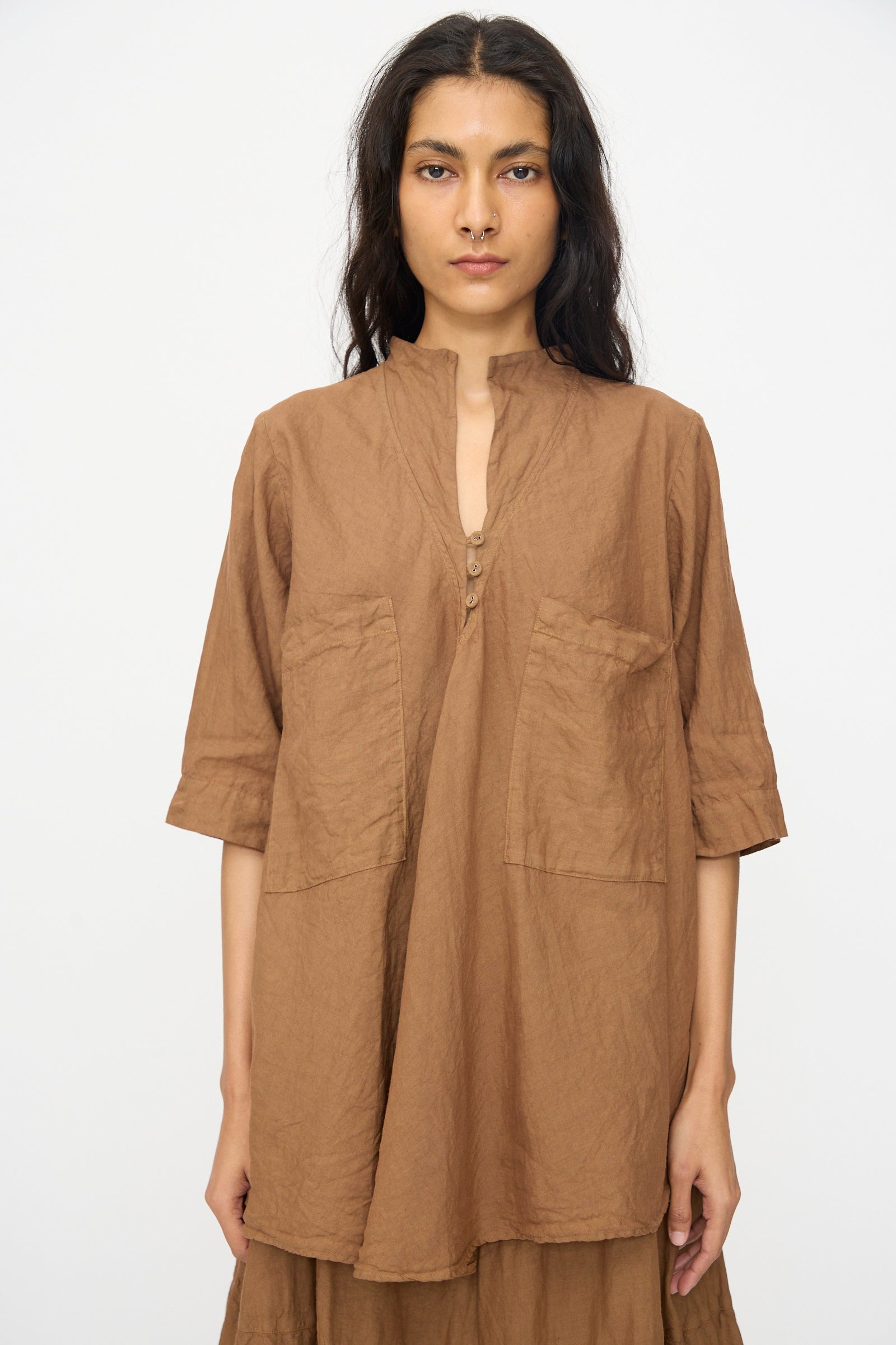 A woman stands against a plain backdrop wearing the Hallelujah Chemise de Garcon Berger in Persimmon Dye—a loose-fitting linen blouse with front buttons and large pockets, made in Japan for stylish comfort.