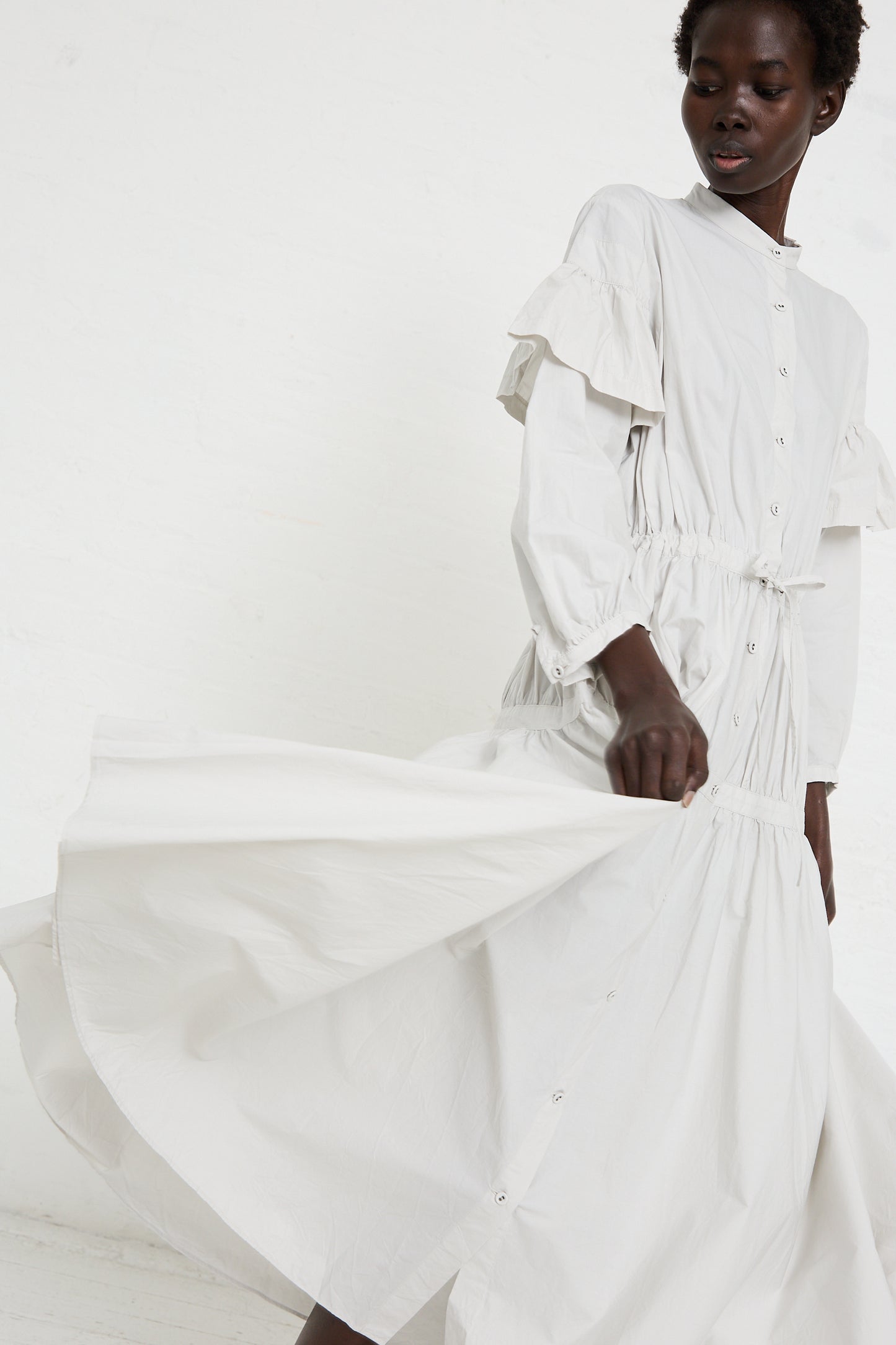 A person wearing a long white Cotton Chemise à la Reine in Turnip with ruffled sleeves and button details by Hallelujah, handmade in Kyoto, poses against a plain white background. The dress flows outward as they extend their arm.