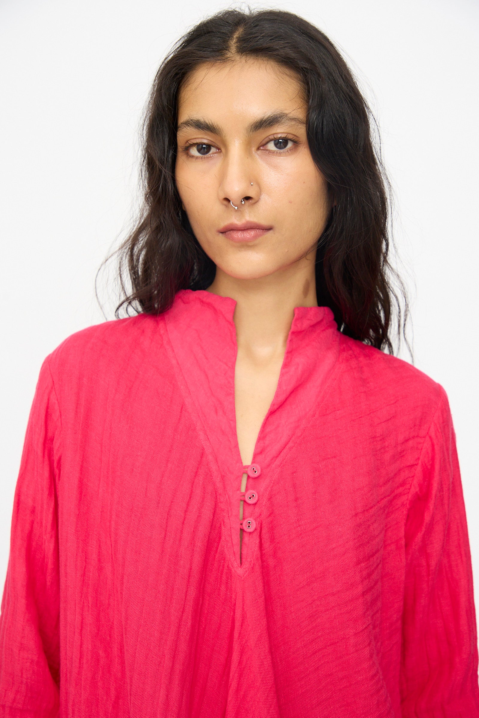 A person with long dark hair and a nose ring stands against a white background, wearing the Hallelujah Cotton Linen Robe de Femme de Chambre in Cherry Red, featuring a short stand collar and buttons.
