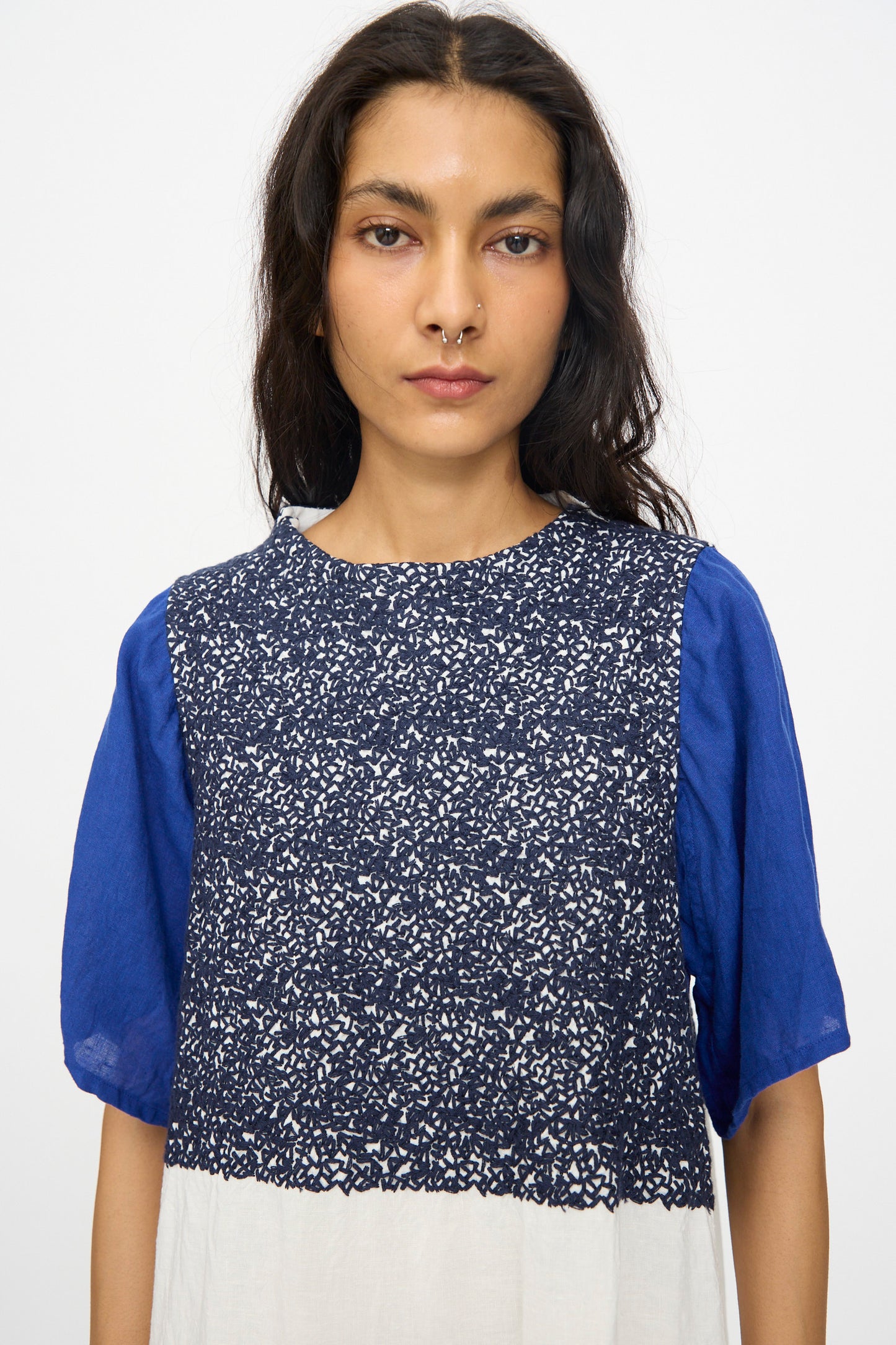 A woman with long dark hair and a nose ring wears the Hallelujah French Linen La Pucelle Dress in Off White Navy Stitch, featuring short sleeves and a blue patterned design, standing against a plain background.