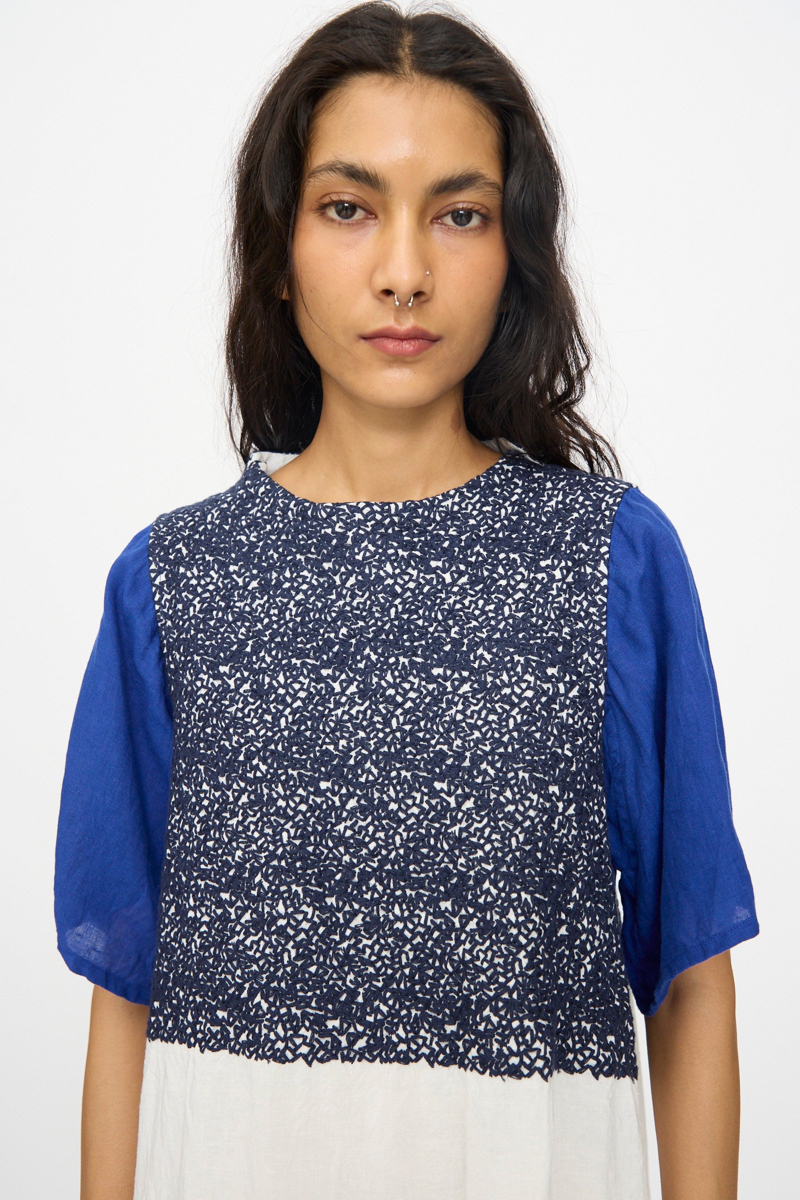 A woman with long dark hair and a nose ring wears the Hallelujah French Linen La Pucelle Dress in Off White Navy Stitch, featuring short sleeves and a blue patterned design, standing against a plain background.