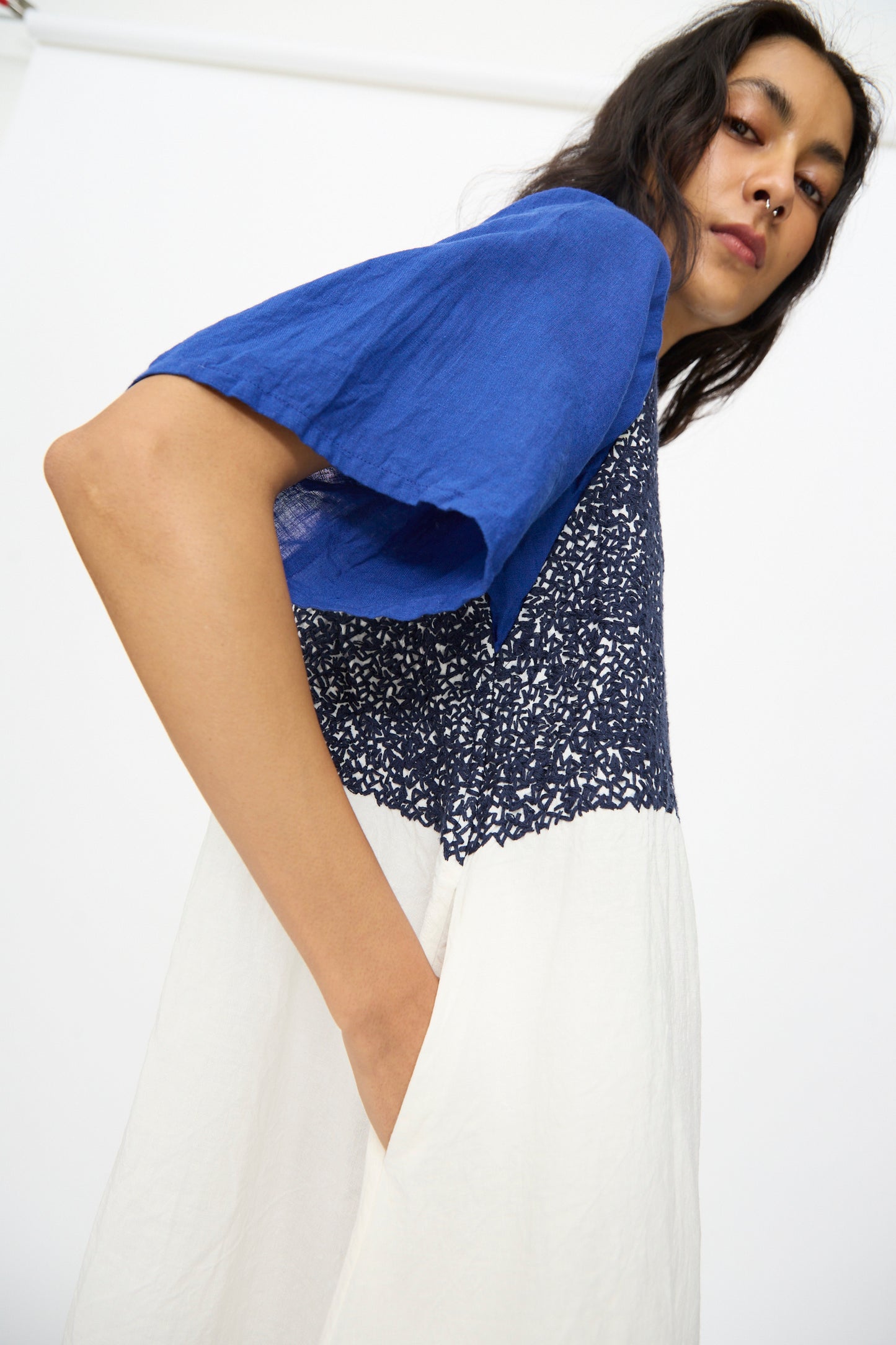 A person stands with one hand in their pocket, wearing the Hallelujah French Linen La Pucelle Dress in Off White Navy Stitch. The short-sleeve midi dress is shown from a low angle against a plain white background.