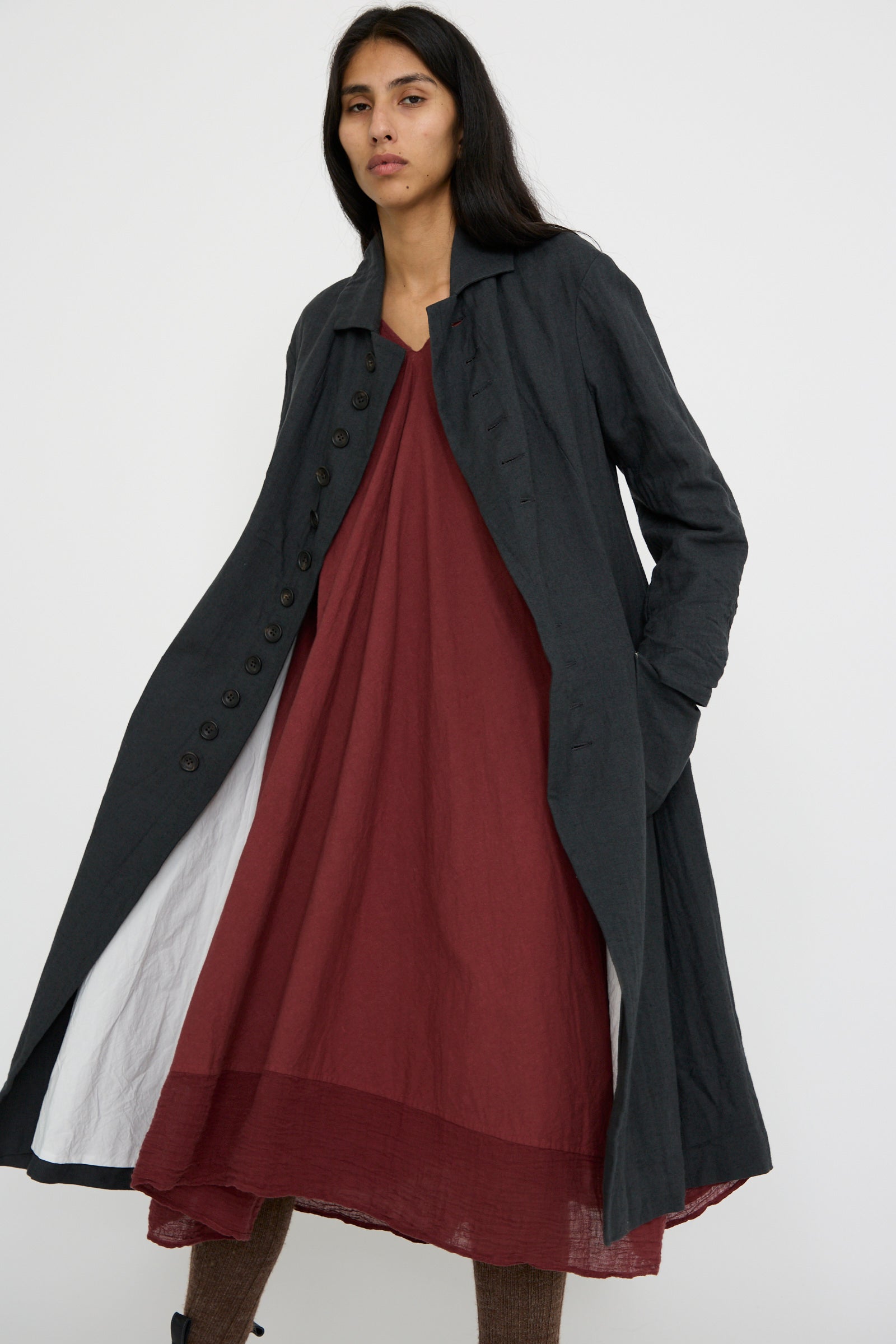 A person wearing the Herringbone Early 20th c. Manteau in Gray by Hallelujah, featuring a classic fold-over collar, draped over a maroon dress, stands against a white background.