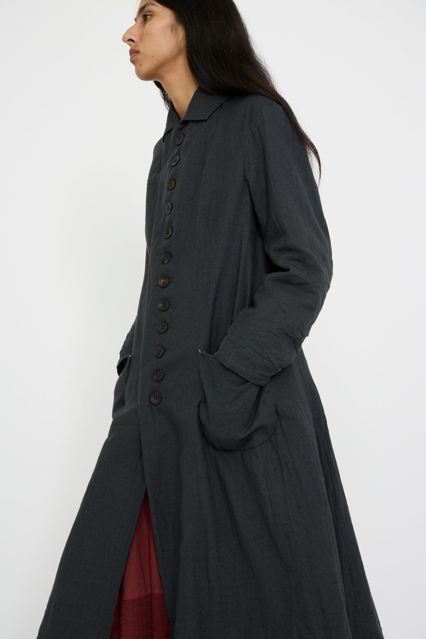 A person stands against a plain background, wearing the Herringbone Early 20th c. Manteau in Gray by Hallelujah—a long, dark buttoned coat featuring a classic fold-over collar and hands tucked in pockets.