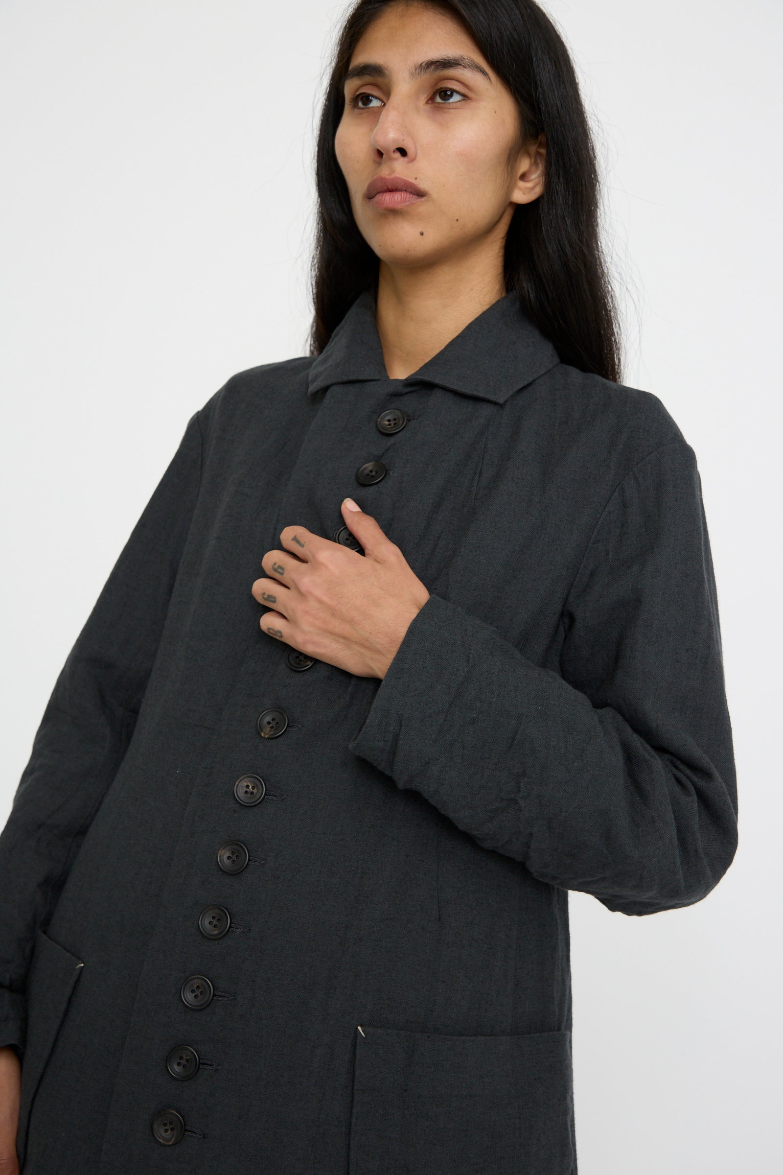 A person stands with one arm on their chest against a plain background, wearing the Herringbone Early 20th c. Manteau in Gray by Hallelujah, featuring a dark buttoned coat with a classic fold-over collar.